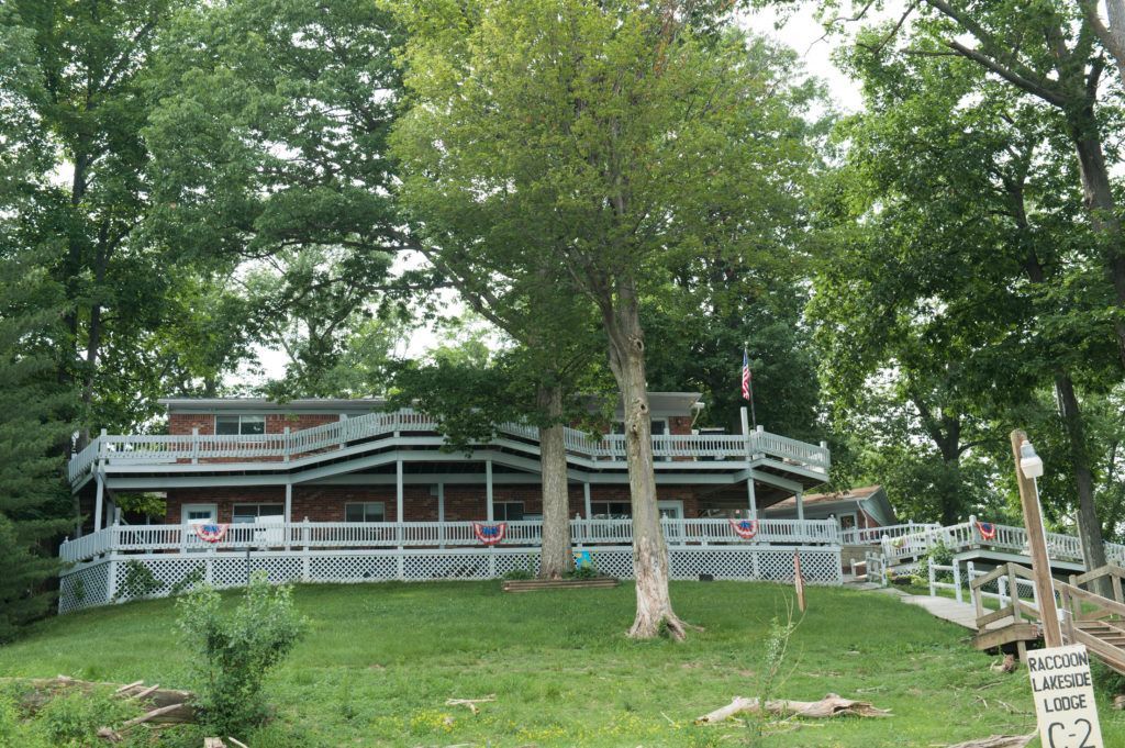 Rustic house with wraparound porch on a grassy hill, surrounded by trees.