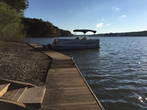 Wooden dock on a lake with a pontoon boat under a blue sky, surrounded by trees and shoreline.