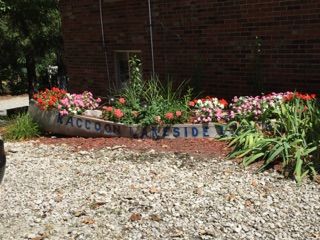 Flower bed with “Welcome” sign beside a brick building and gravel path