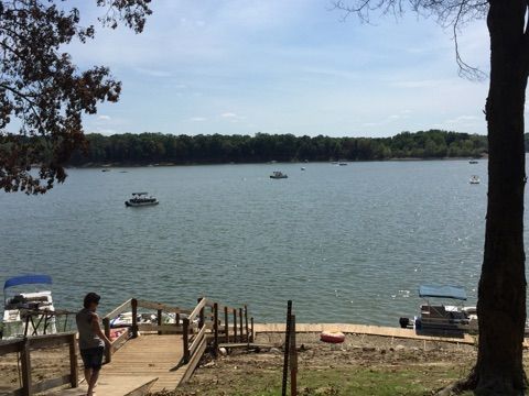 Lake view from a dock with a person standing by wooden stairs, boats on calm water, and trees framing the shore.