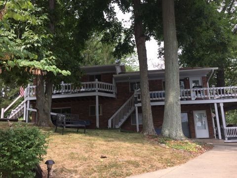 Two-story house on a wooded slope with wraparound porch and stairs leading to the entrance