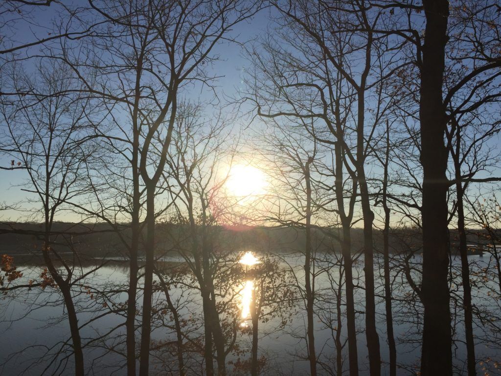 Sunset over a lake, with bare trees silhouetted against the bright sky and reflected light on the water