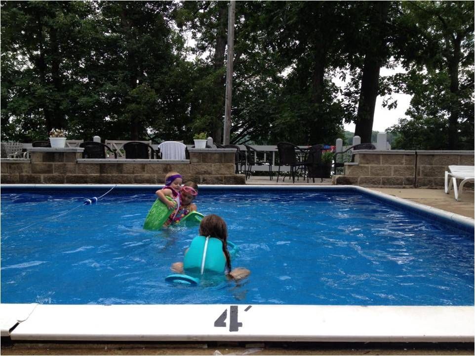 Children in bright swimsuits play in an outdoor swimming pool surrounded by trees.