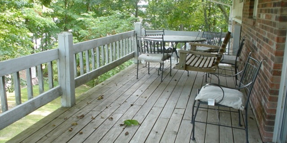 Wooden porch with patio chairs and table beside a brick wall, overlooking green trees and a yard.