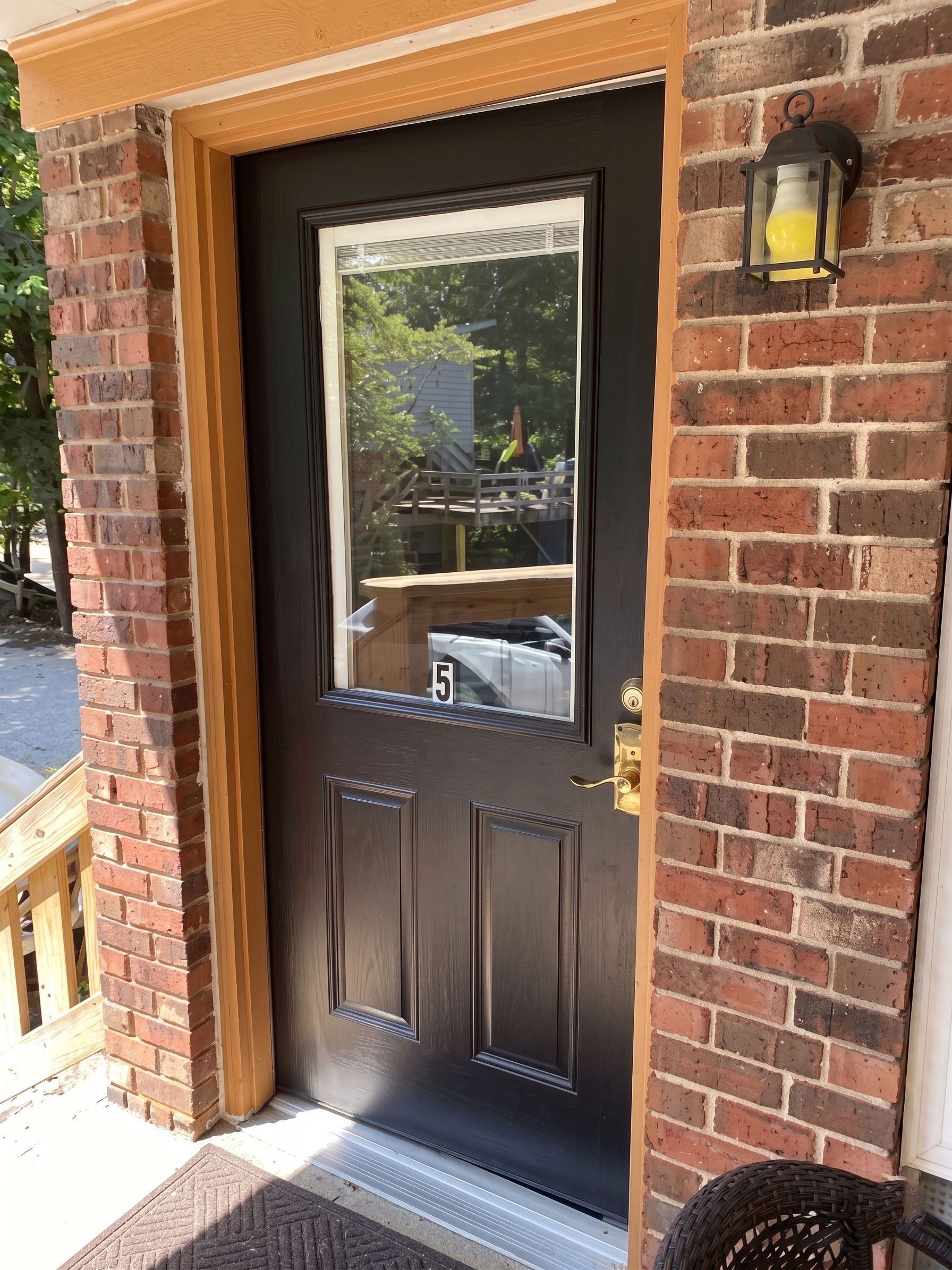 Black front door with window, brass handle, and lantern on a brick house porch.
