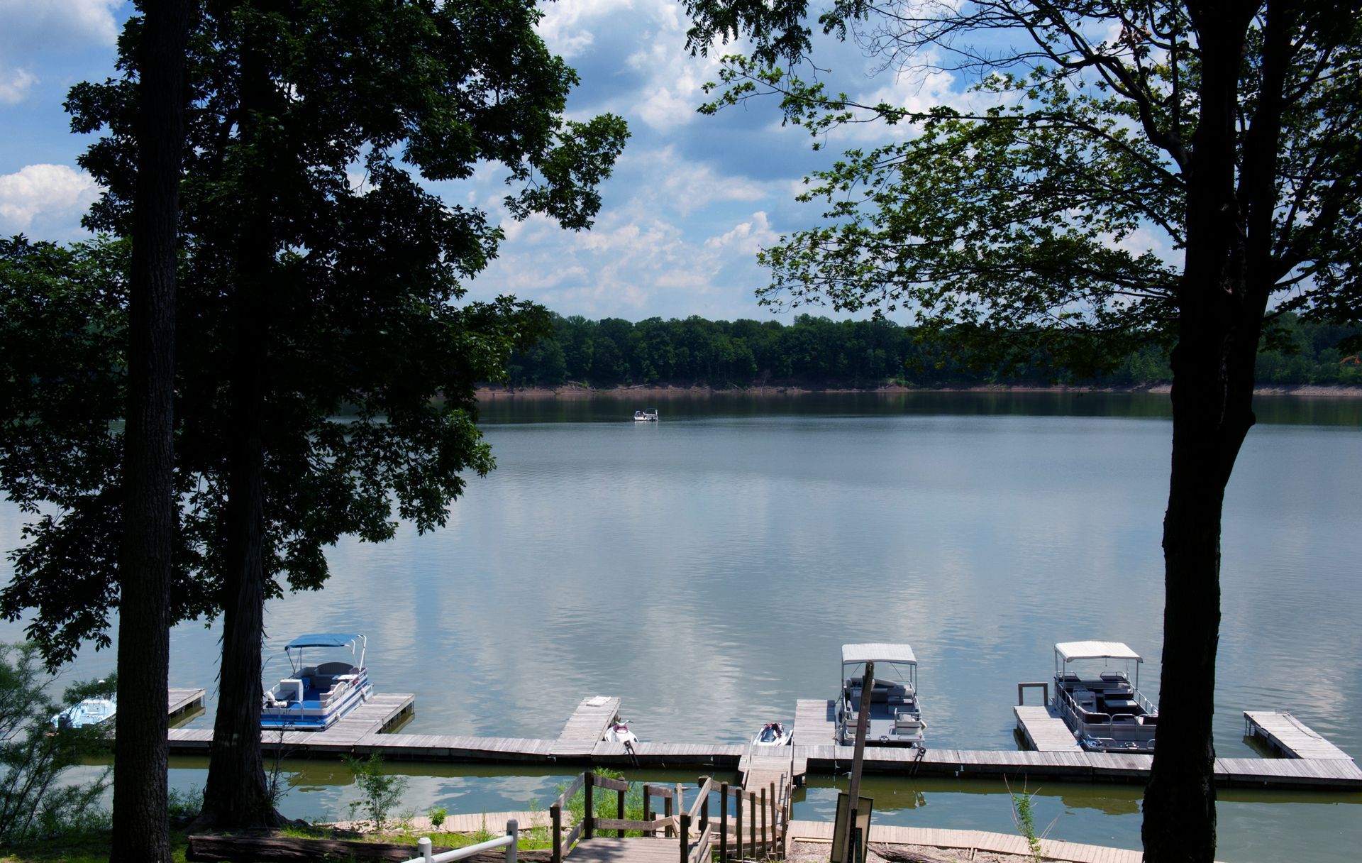 Lake view from a shaded dock with boats, trees, and calm water under a partly cloudy sky