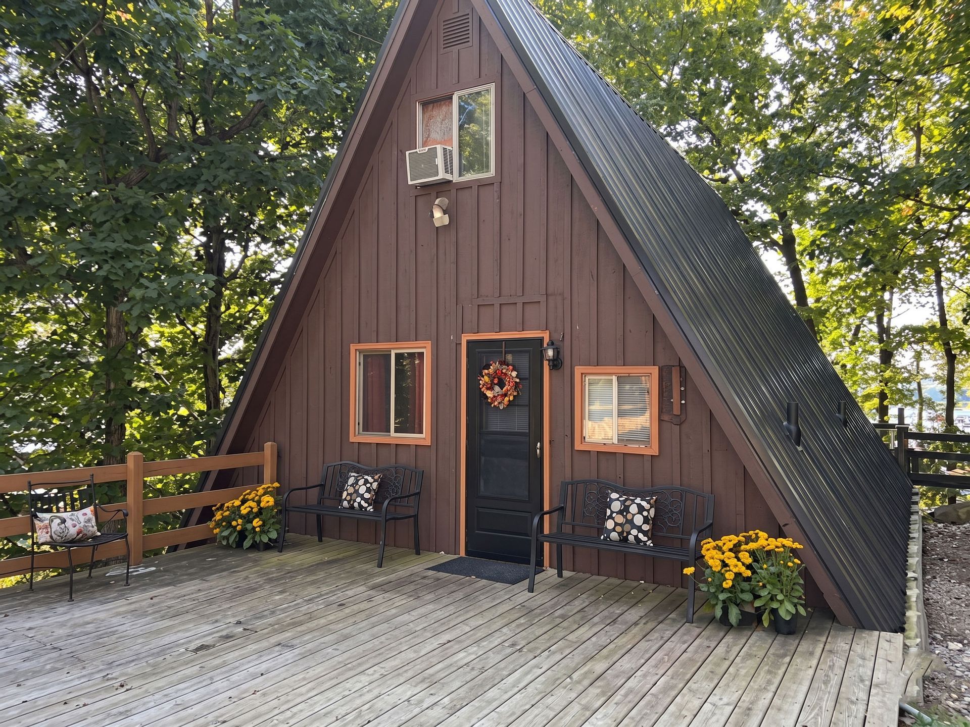 A small brown A-frame cabin with a black door and flower pots on a wooden deck, surrounded by trees.