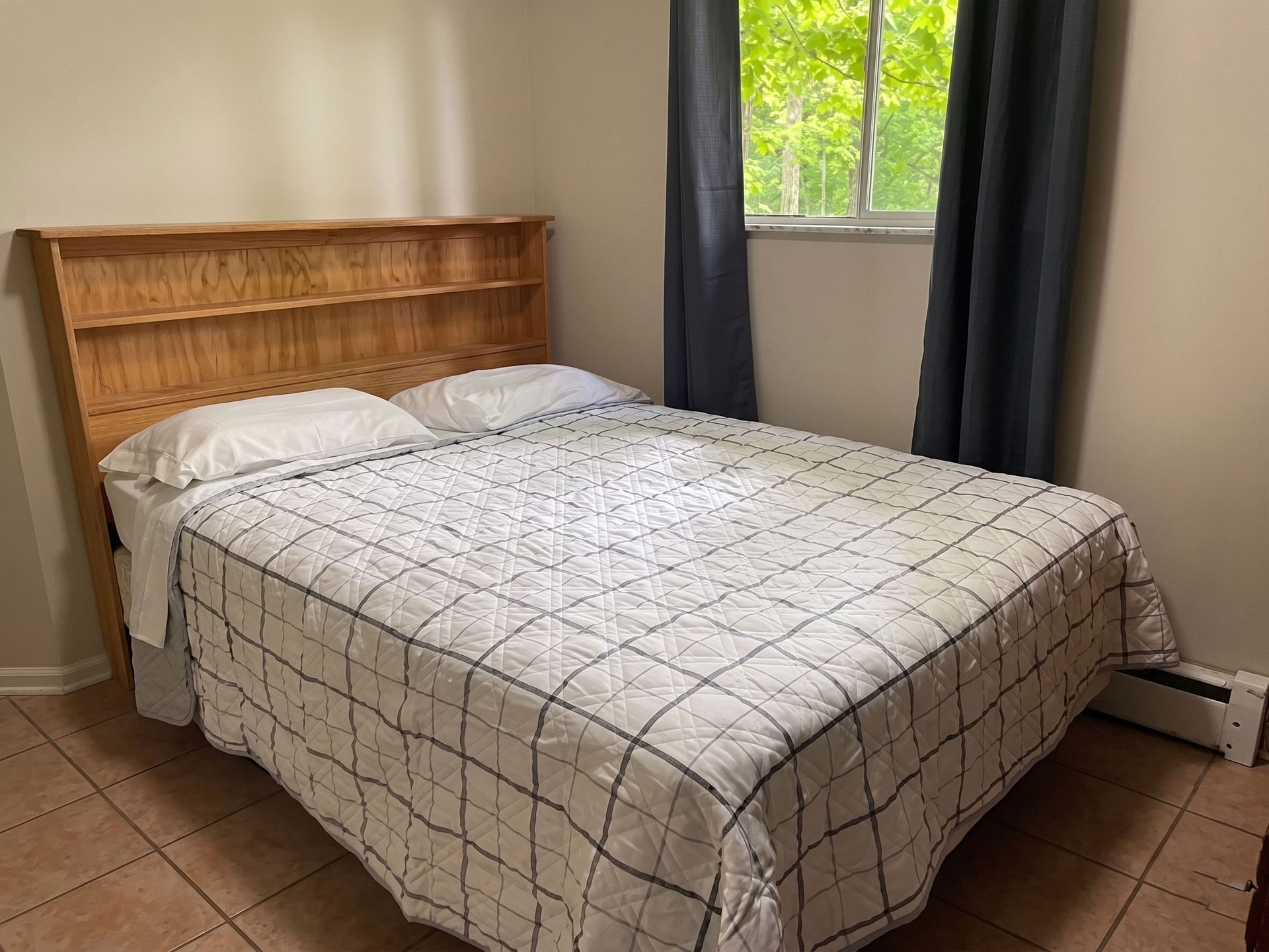 Small bedroom with a bed, wooden headboard, tiled floor, and window with dark curtains.