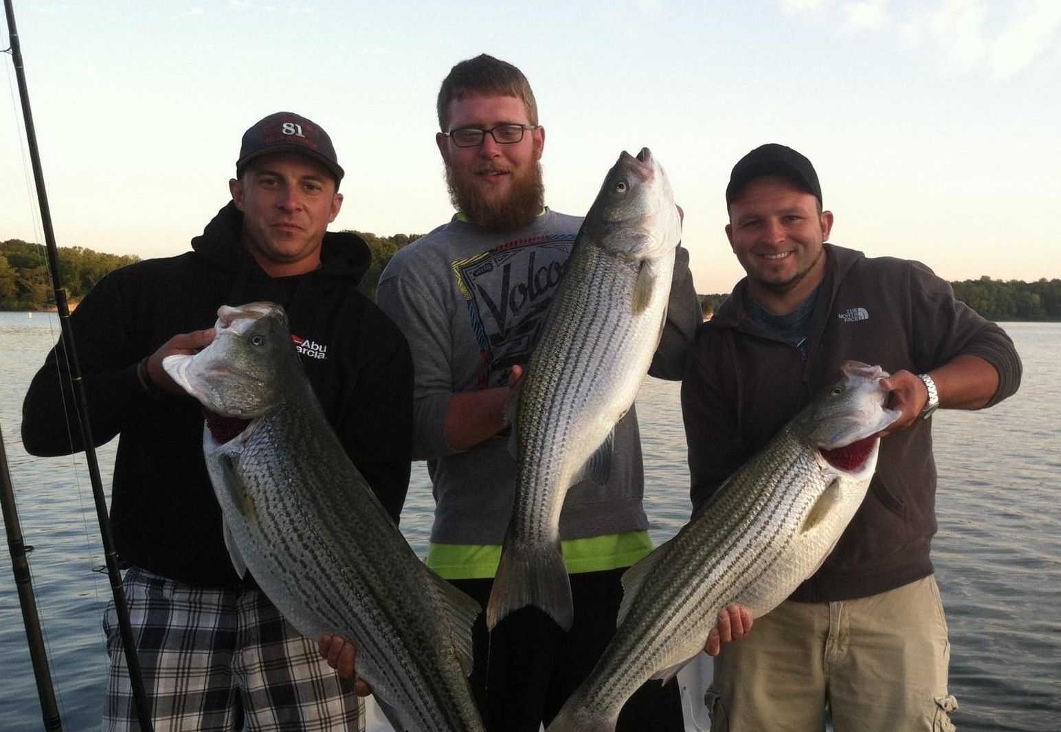 Three men holding large fish on a boat at sunset.