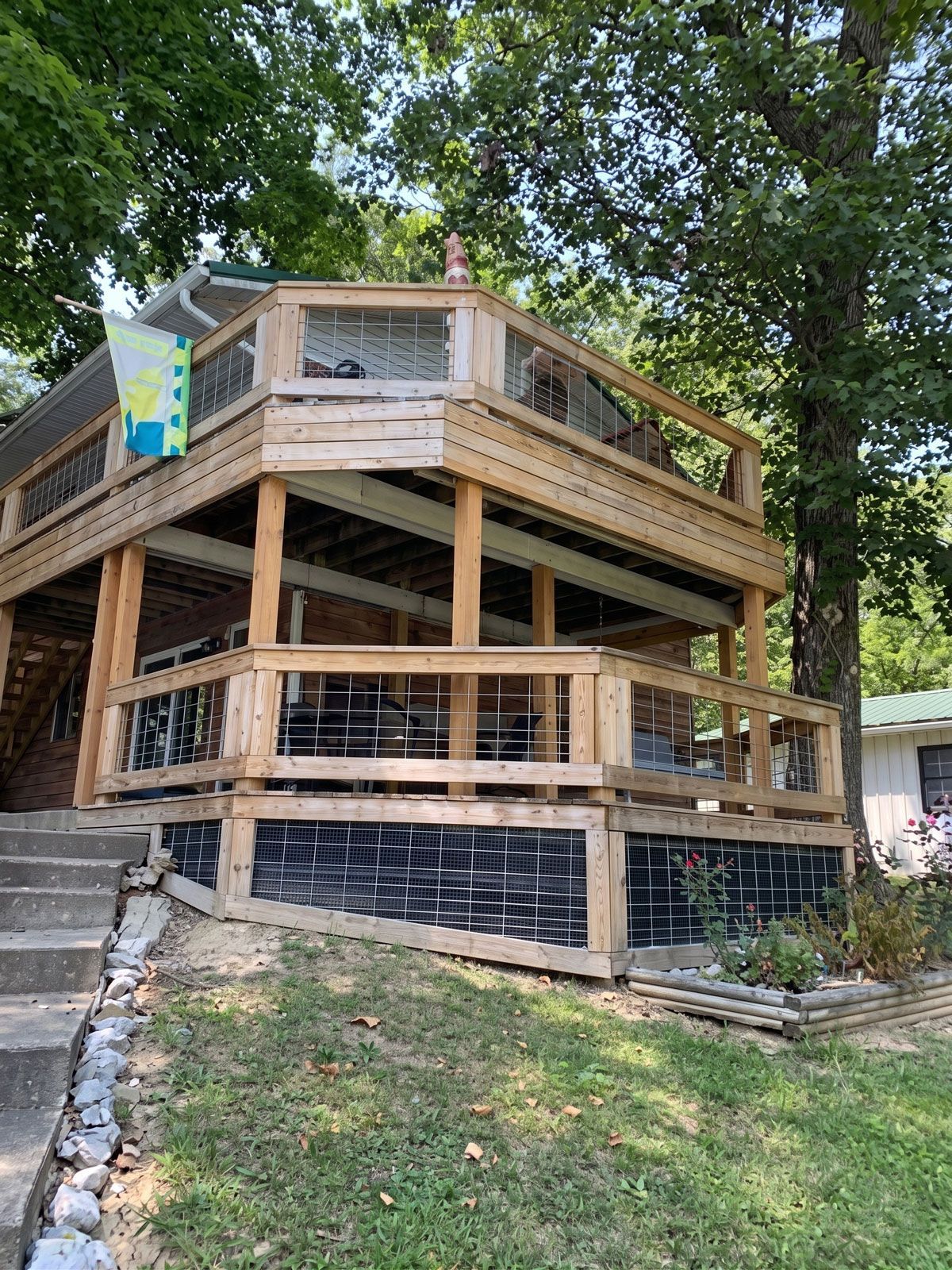Wooden two-story deck and porch structure on a sloped yard, surrounded by trees and railings.