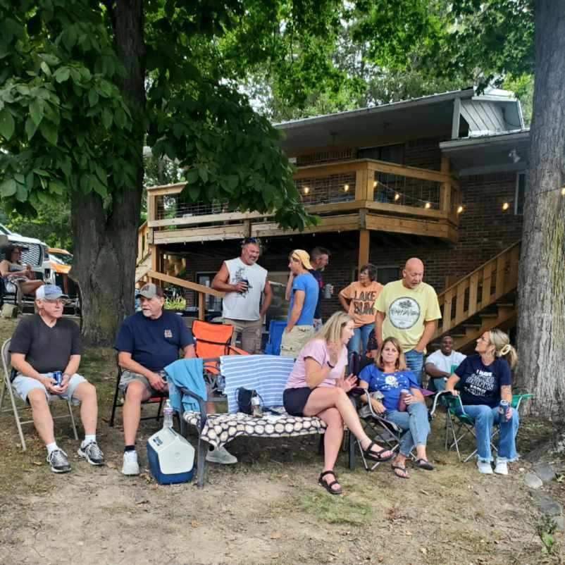 Group of people sitting and standing outside a rustic cabin under trees, relaxing together at a picnic table