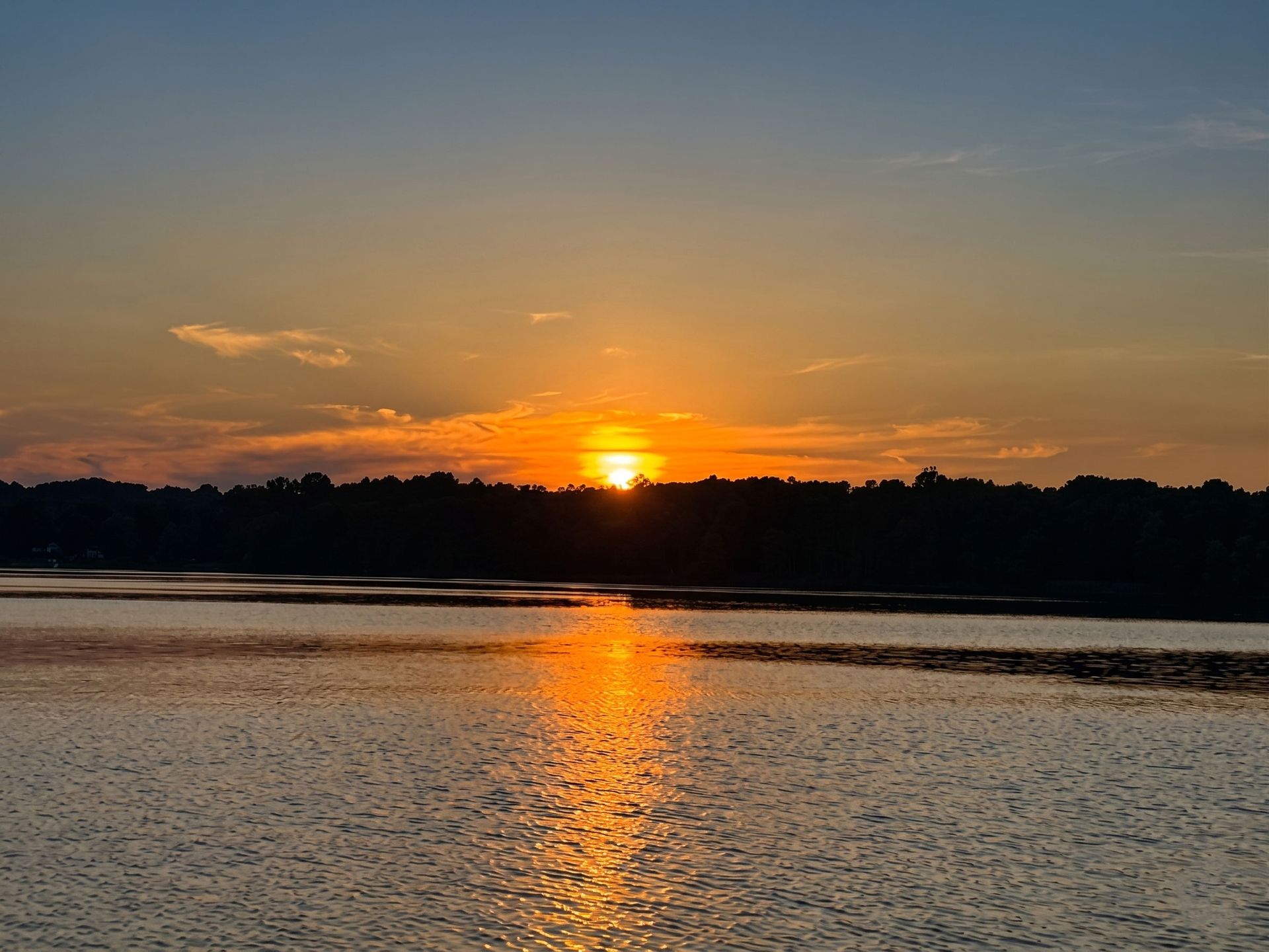 Sunset over a calm lake, with orange reflections on the water and a dark treeline silhouette