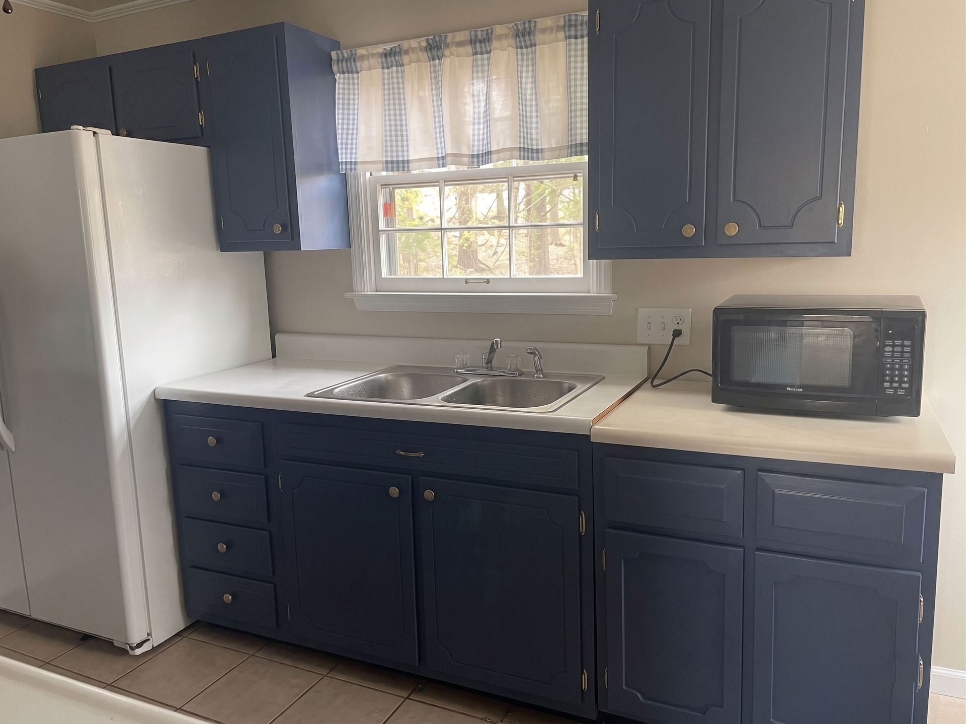 Small kitchen with dark blue cabinets, white refrigerator, sink under window, and microwave on counter.