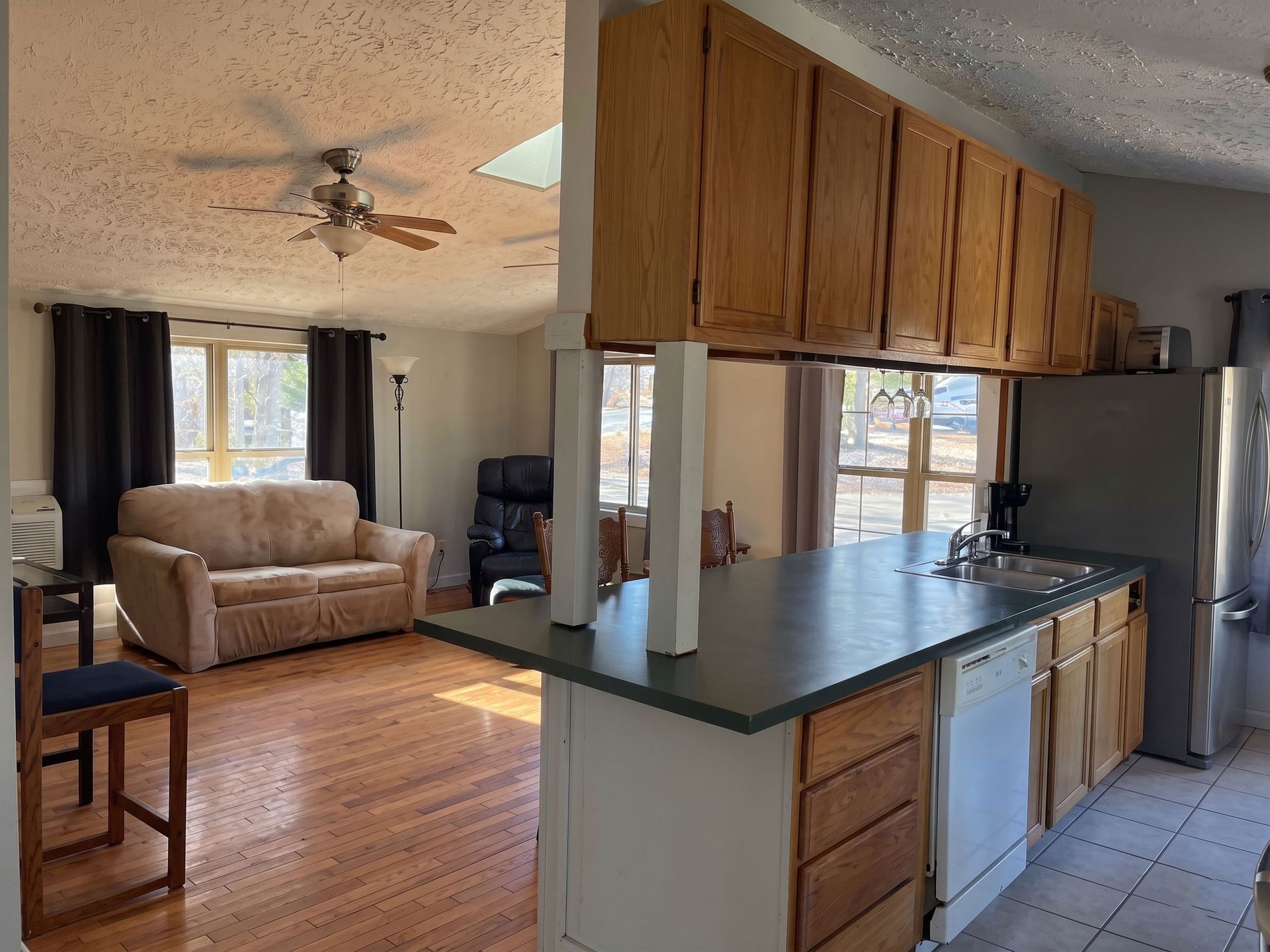 Open kitchen with wood cabinets and dark counters overlooking a living room with beige sofa and ceiling fan.