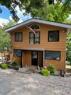 Two-story tan house with black windows, front porch, and gravel driveway under leafy trees