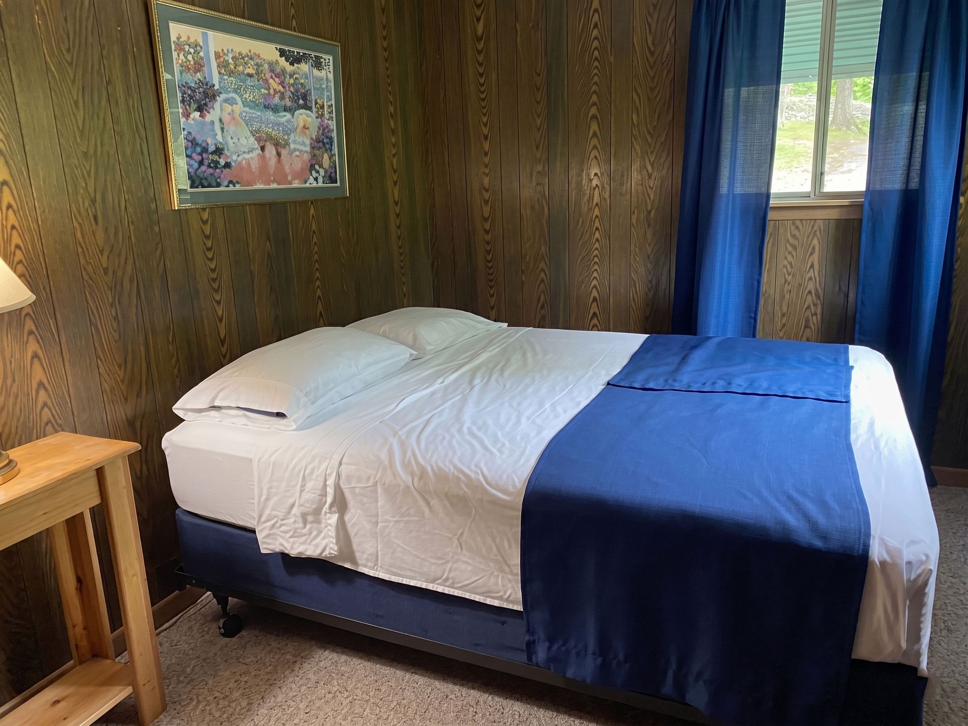Small bedroom with a made bed, wood-paneled walls, blue curtains, and a framed picture above the bed.