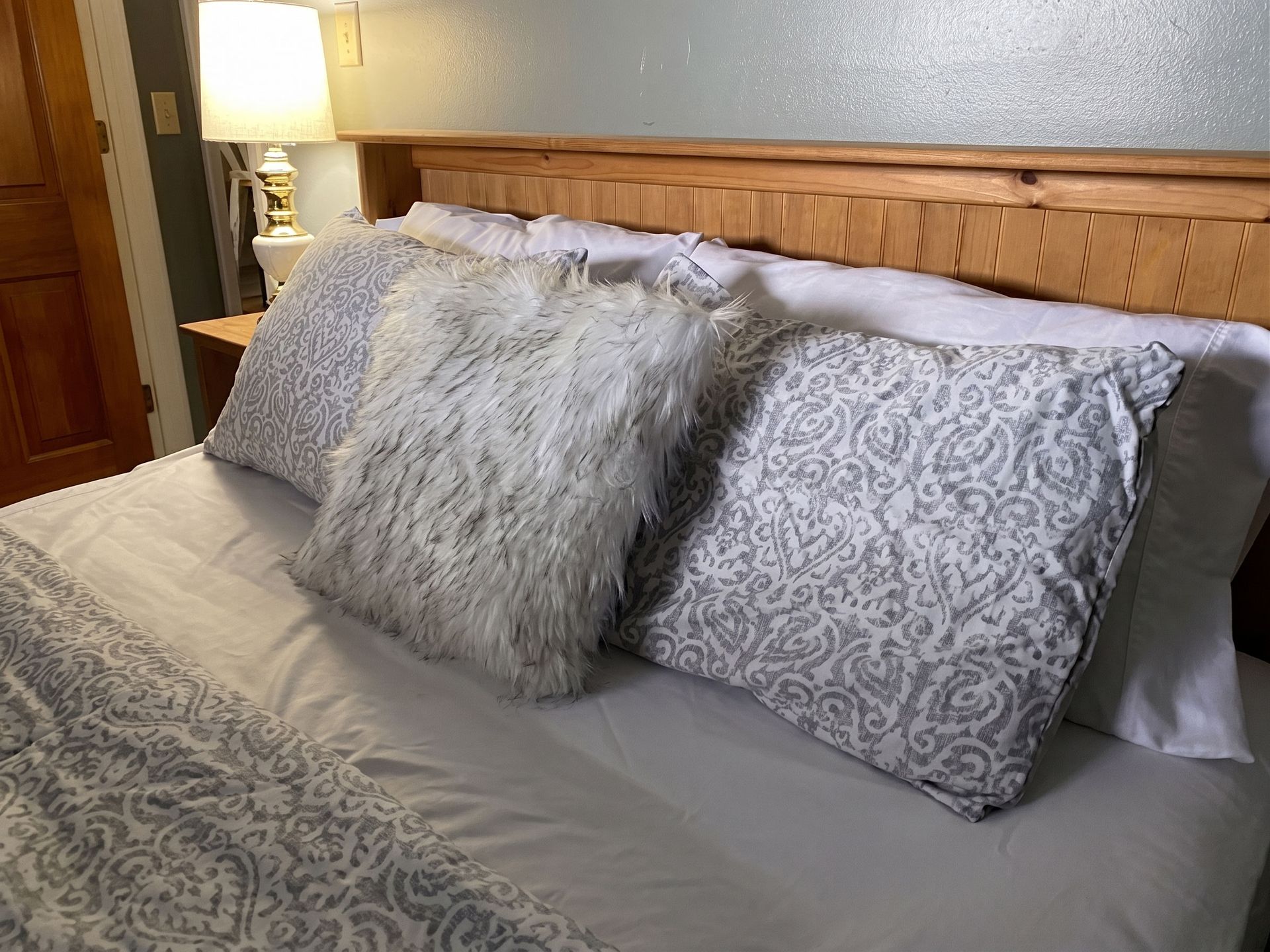 Cozy bed with gray patterned pillows, white bedding, and a wooden headboard beside a lit lamp.
