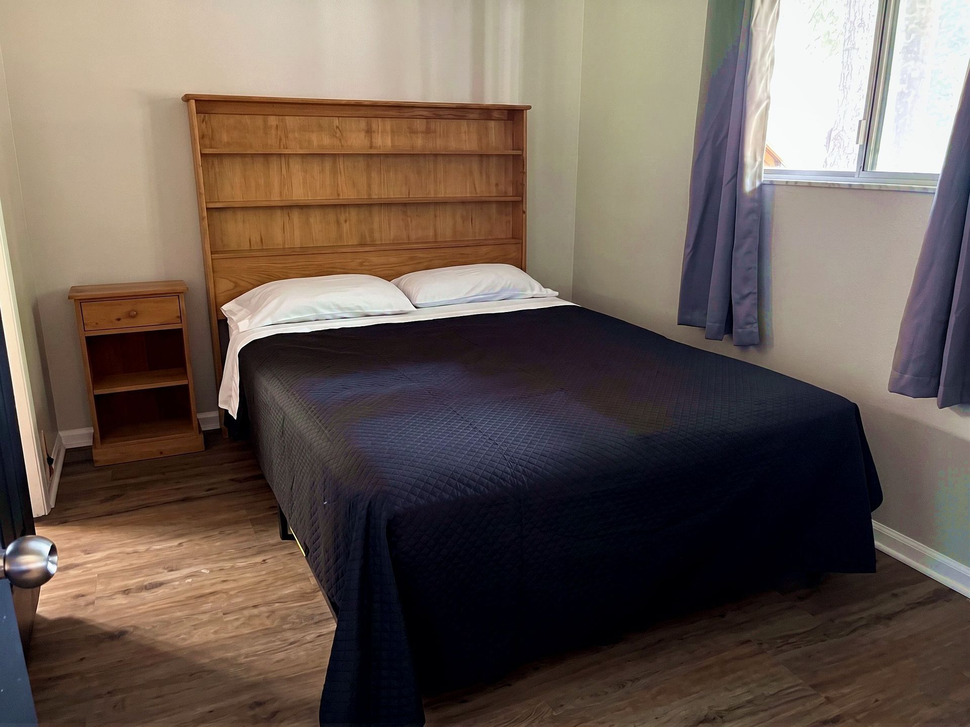 Small bedroom with a double bed, wooden headboard, bedside table, and window with curtains.