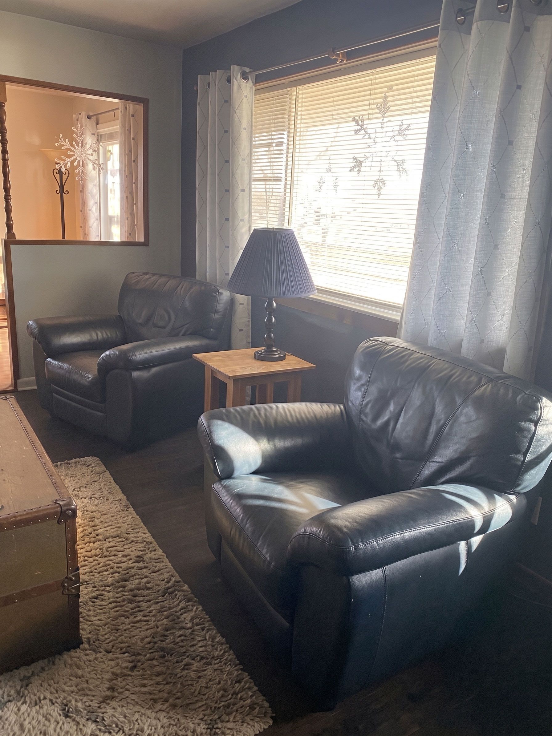 Living room with two black leather chairs, a side table, lamp, mirror, and window with blinds and curtains