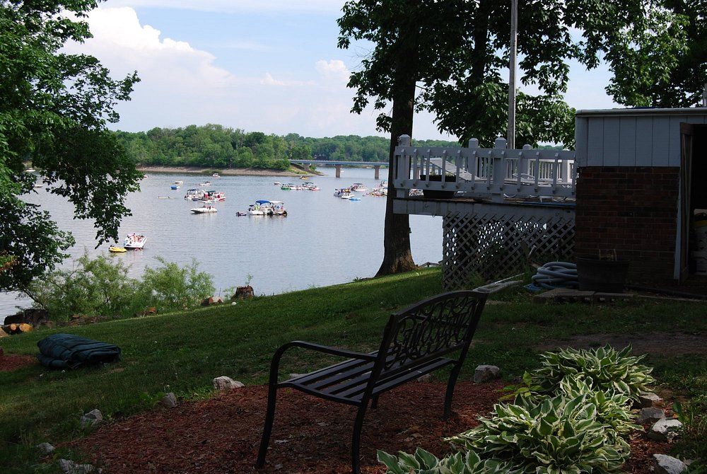 Lakeside dock with boats on calm water, framed by trees and a bench in the foreground.