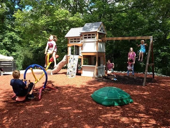 Children playing on a colorful playground with a treehouse, slide, swings, and wood-chip ground