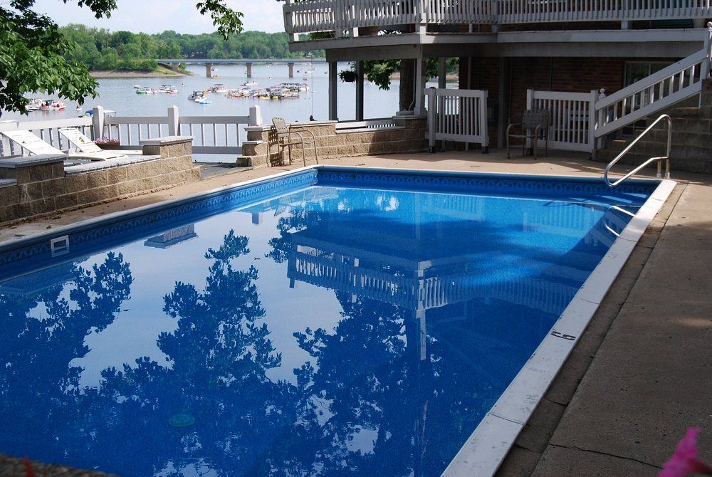 Blue swimming pool beside a lakeside deck and house, with trees reflected in the water
