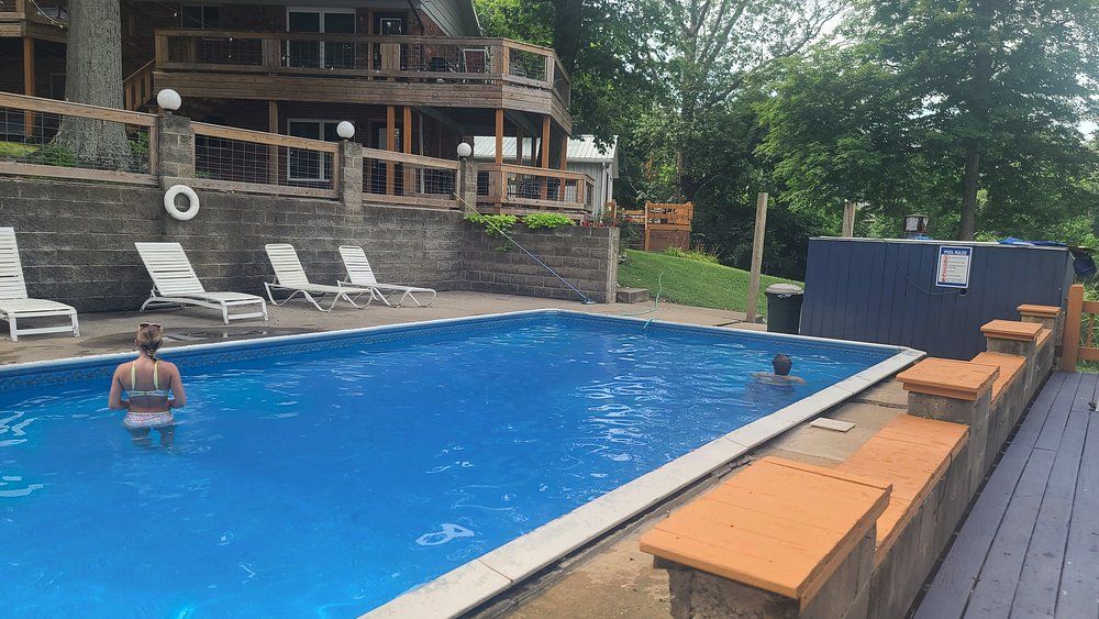 Outdoor pool with a swimmer, lounge chairs, and wooden deck beside a building and trees.