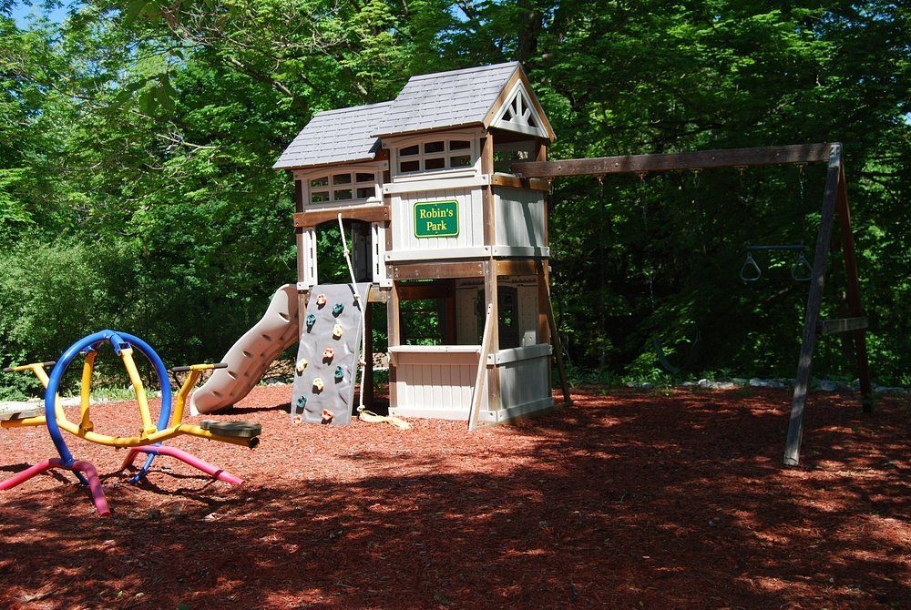 Children’s playground with wooden playhouse, slide, climbing wall, and swings under trees