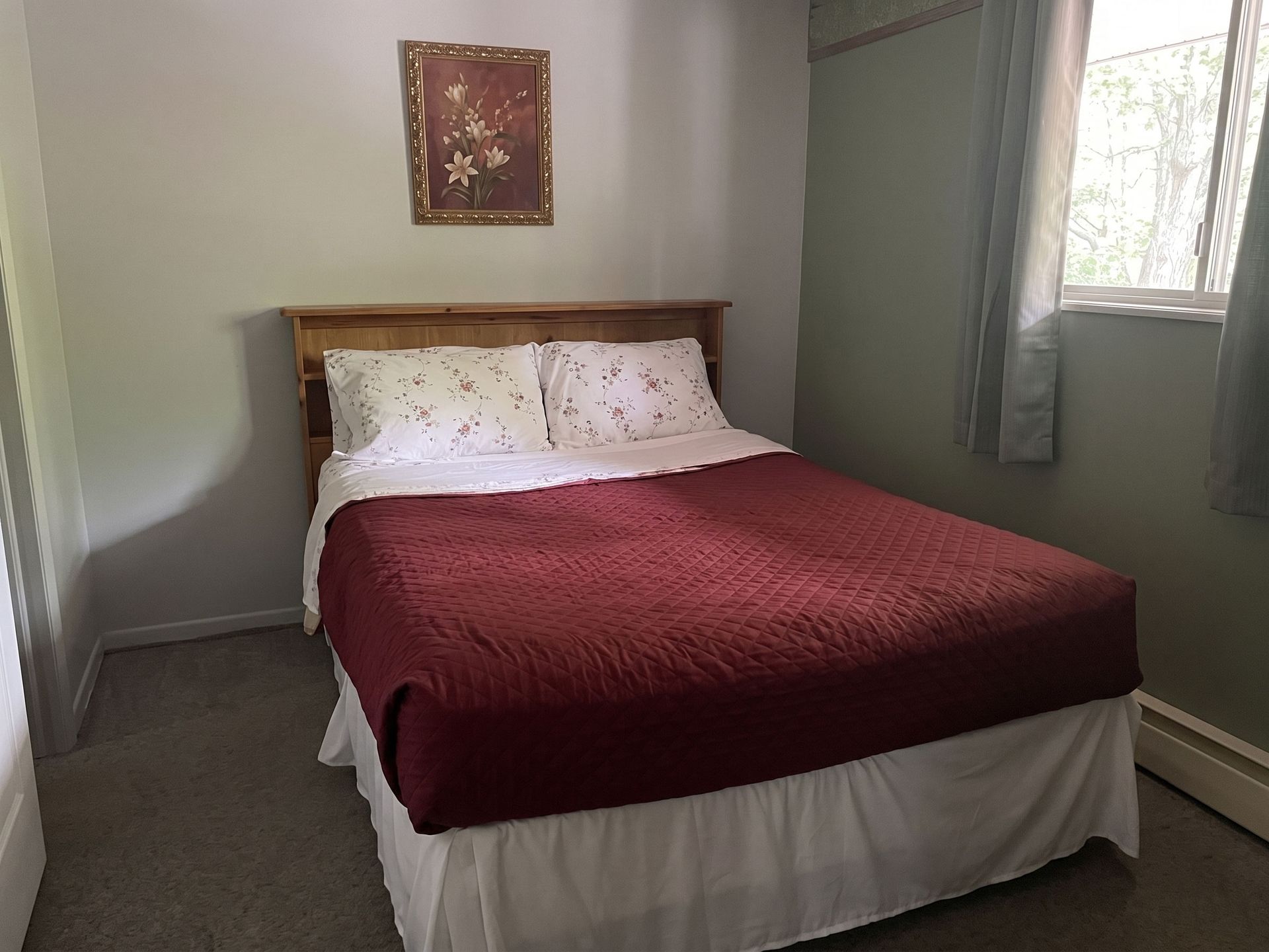 Small bedroom with a bed, burgundy quilt, white bedding, and a window with light green curtains.