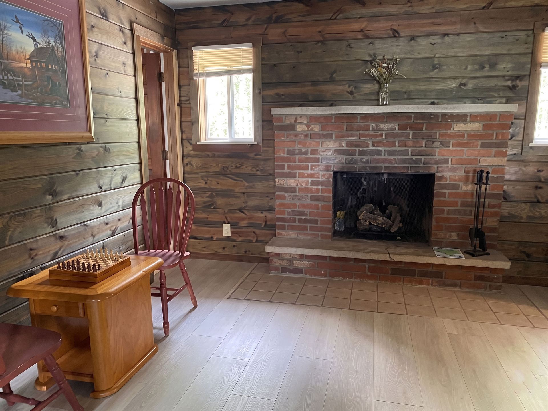 Rustic cabin living room with brick fireplace, wood-paneled walls, chair, and small table with chessboard