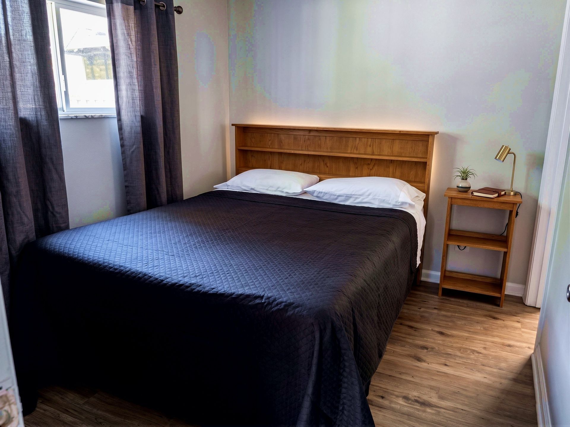 Small bedroom with a black bedspread, wooden headboard, nightstand, and window with curtains