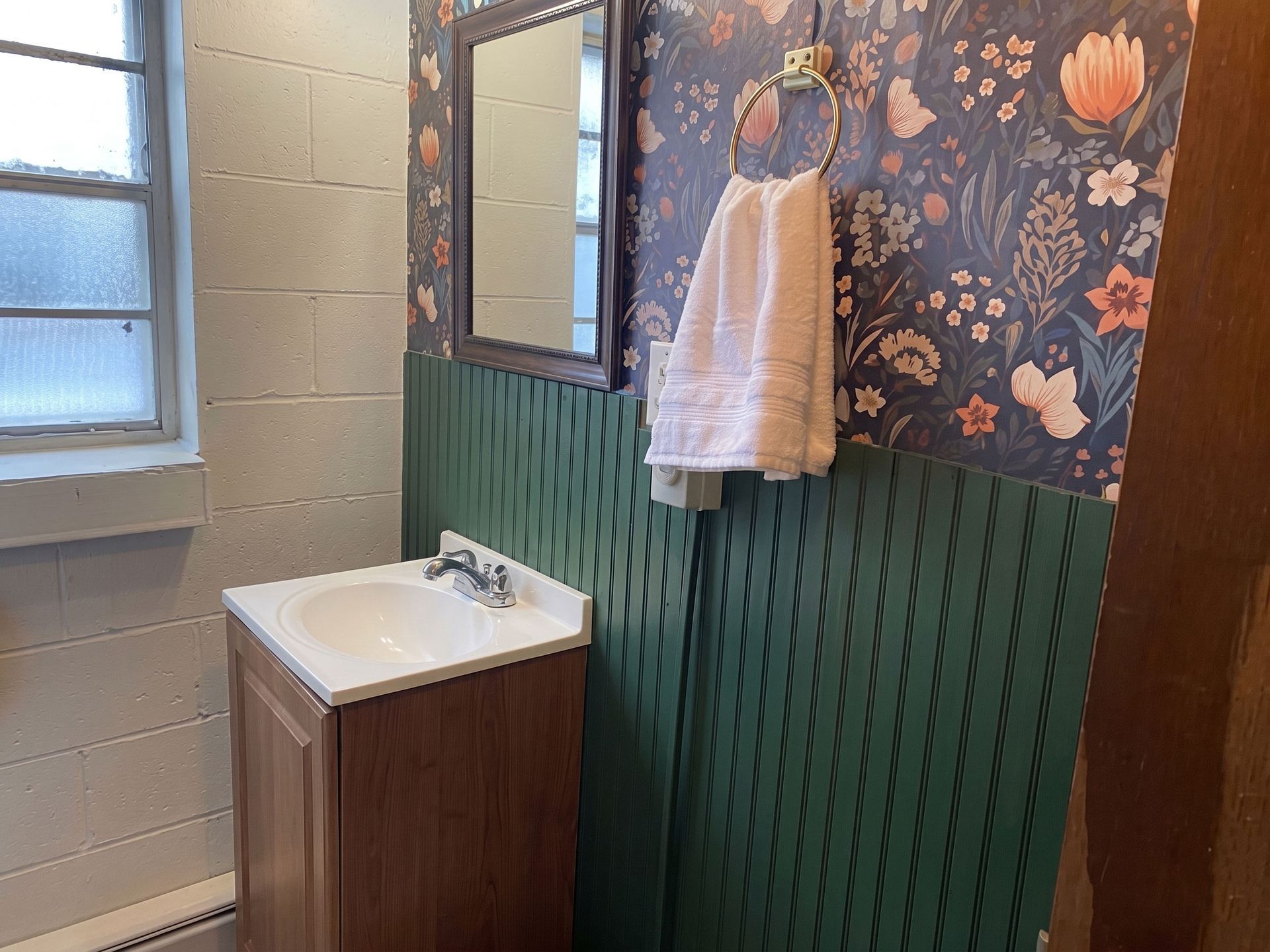 Small bathroom with wood vanity, wall mirror, towel rack, floral wallpaper, and window.