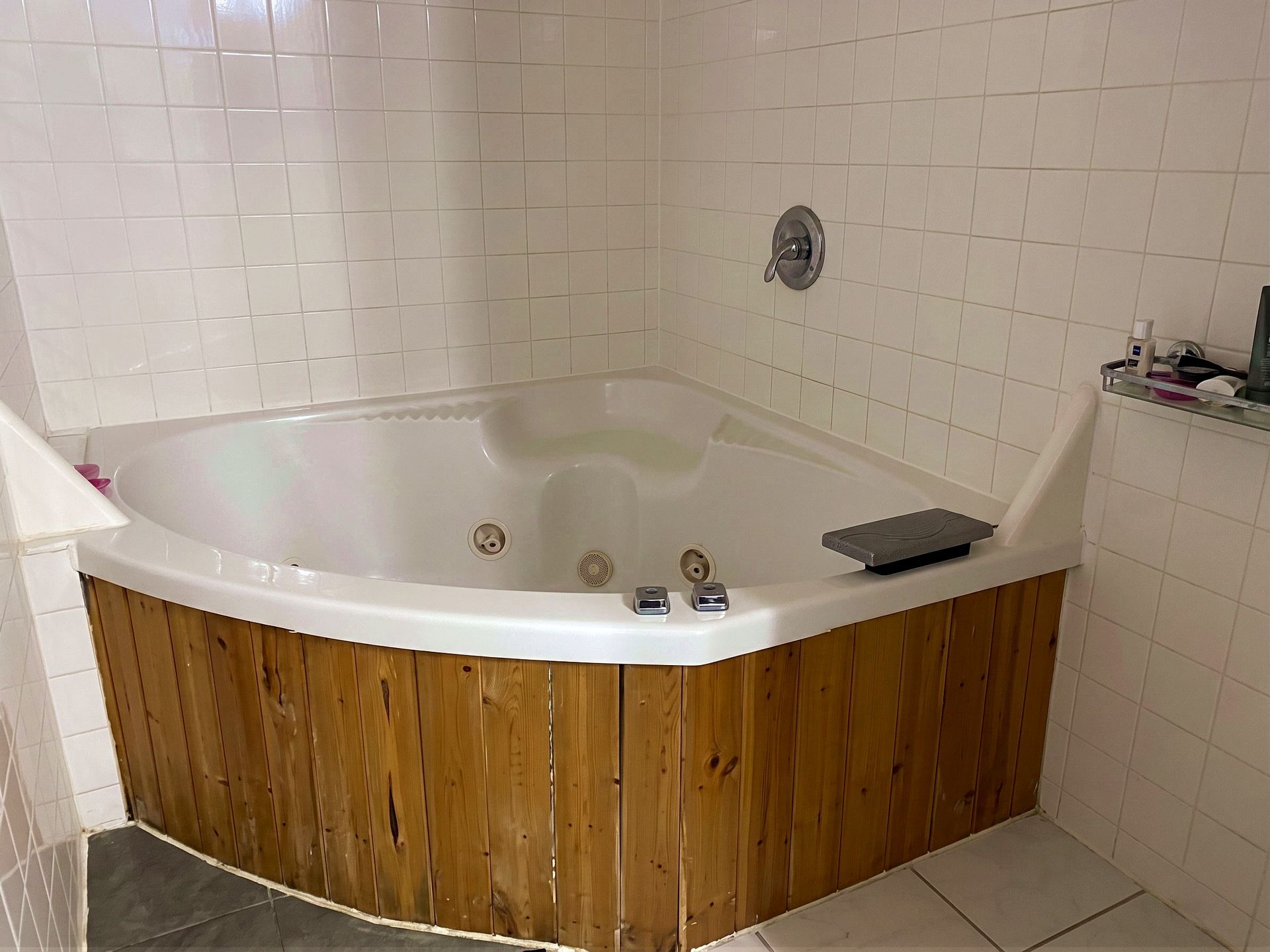 Corner bathtub with wood-paneled front in a white tiled bathroom