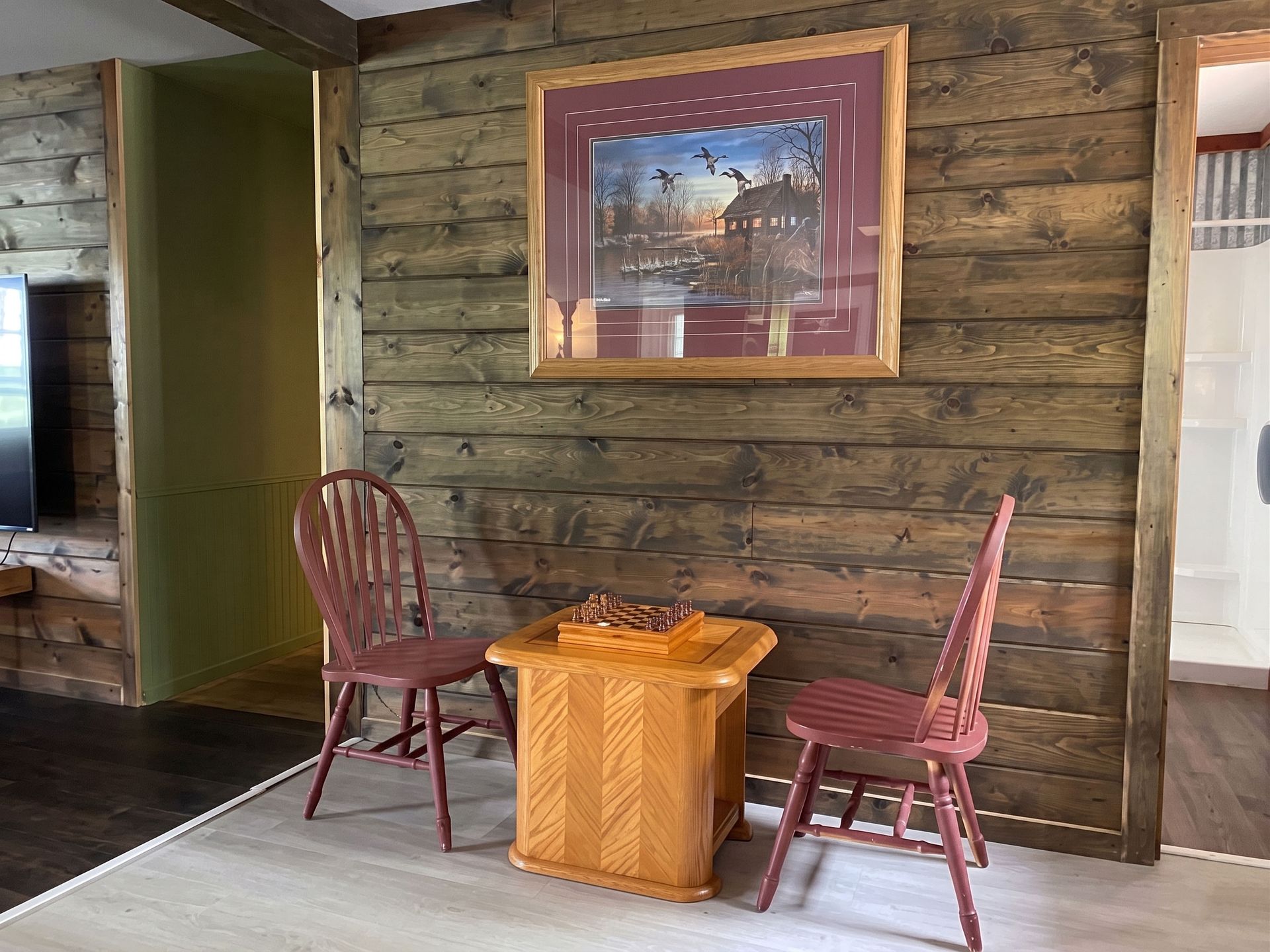 Wood-paneled porch with two red chairs, a small table, and a framed painting on the wall
