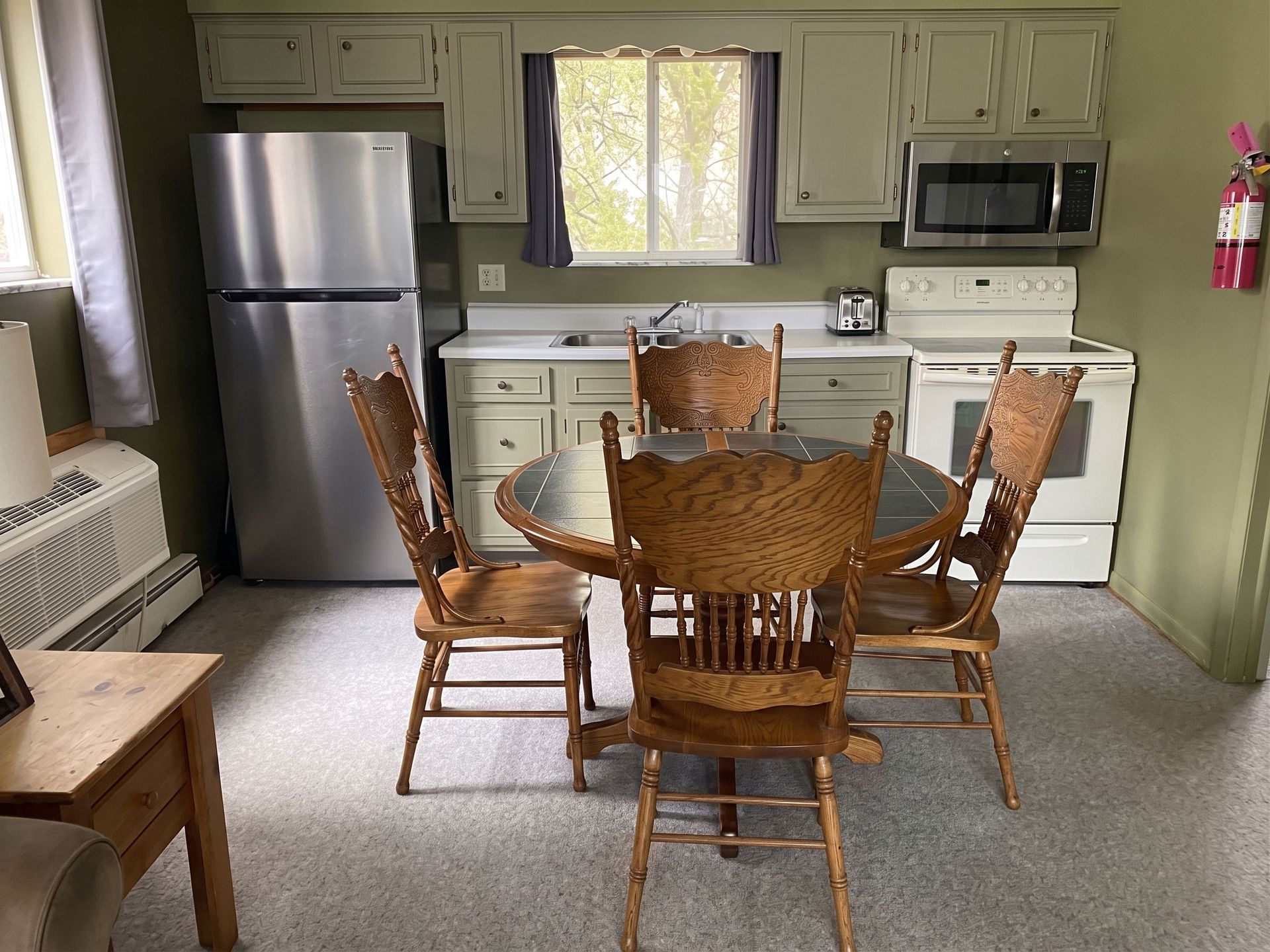 Small kitchen with stainless fridge, white stove, wooden dining table and chairs, and a window over the sink.