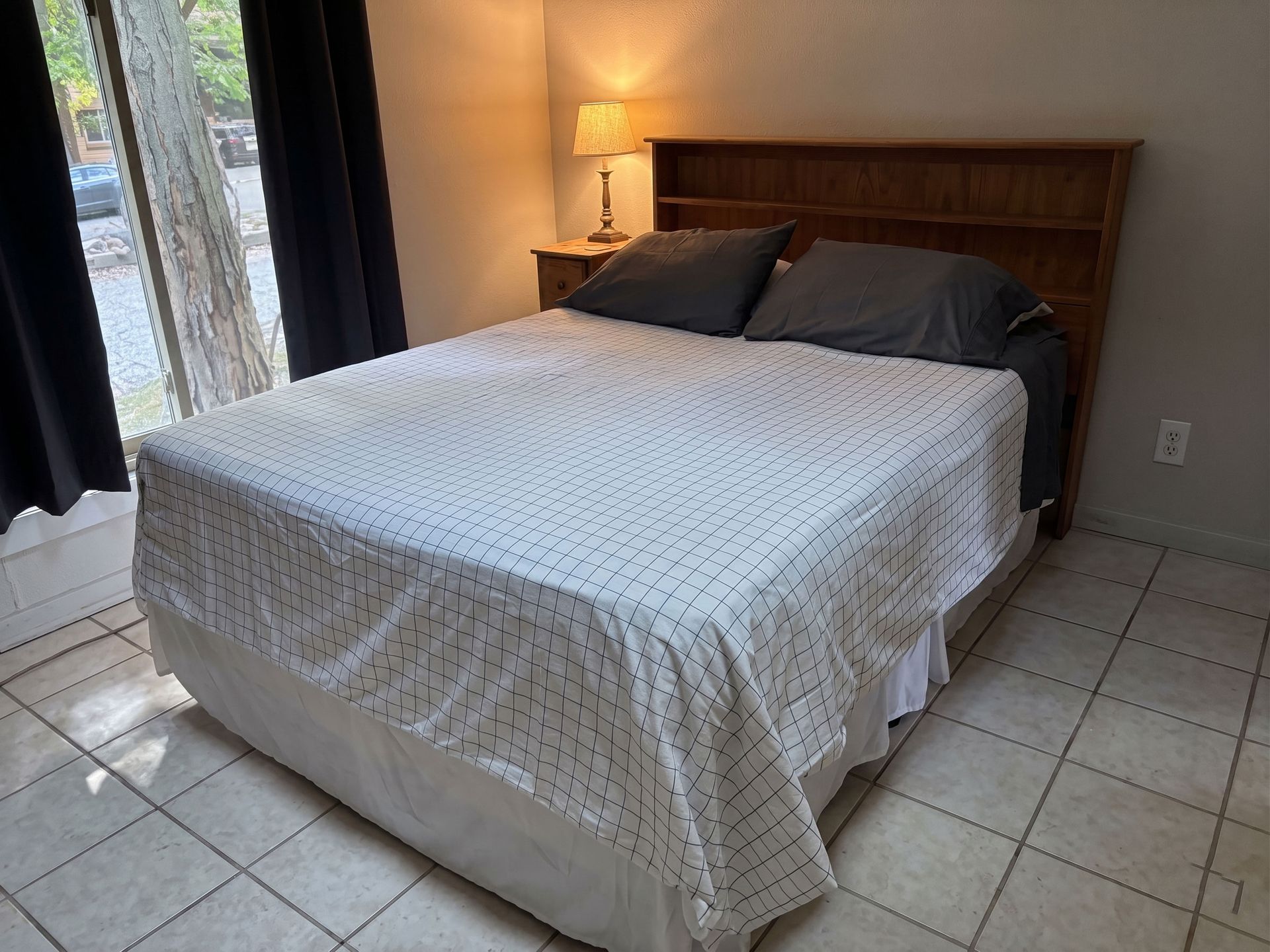 Bedroom with a made bed, wooden headboard, bedside lamp, and tiled floor by a window and curtain.