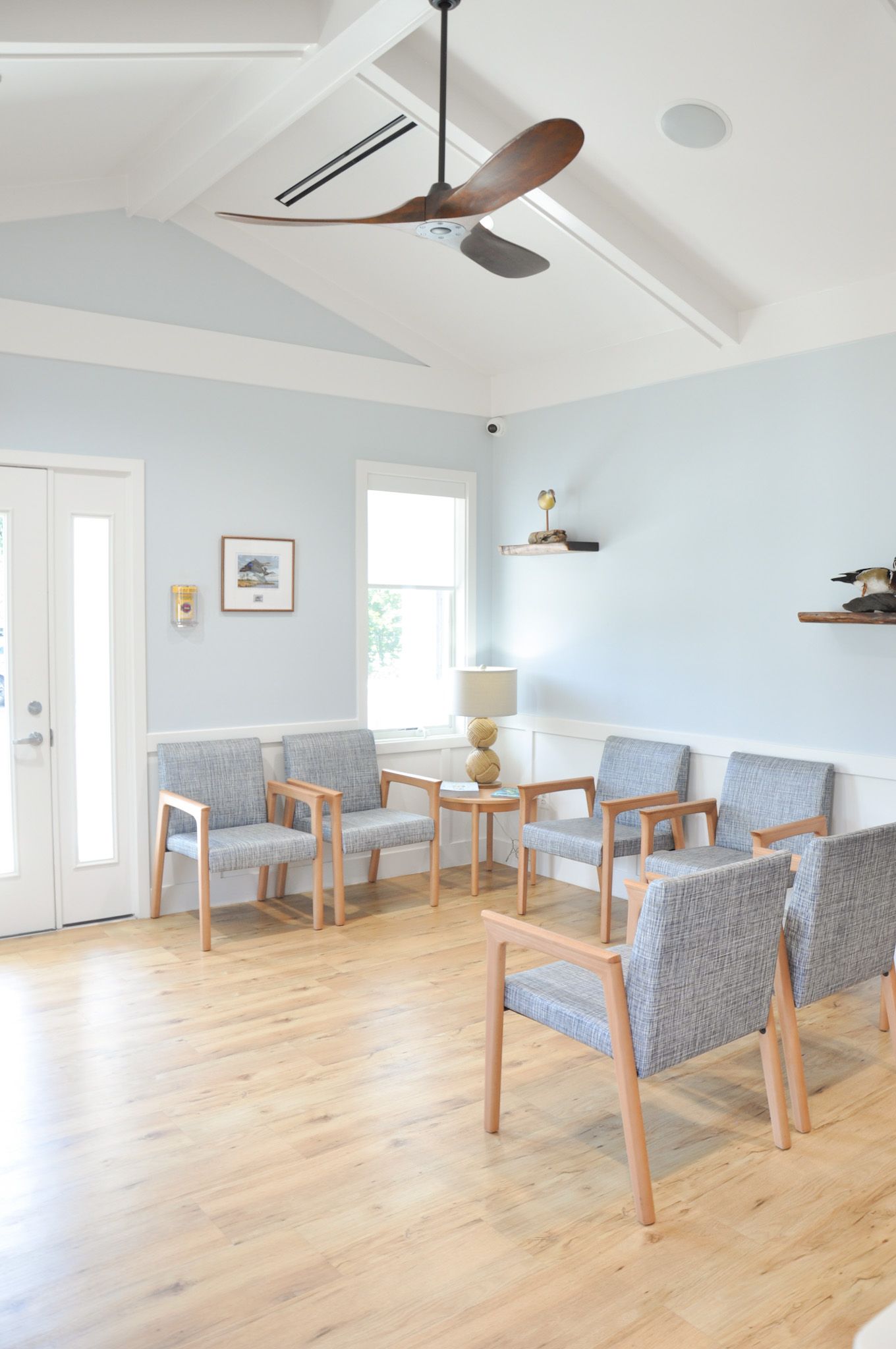 Waiting room with light blue walls, wood floors, and gray chairs. A ceiling fan and small table are present.