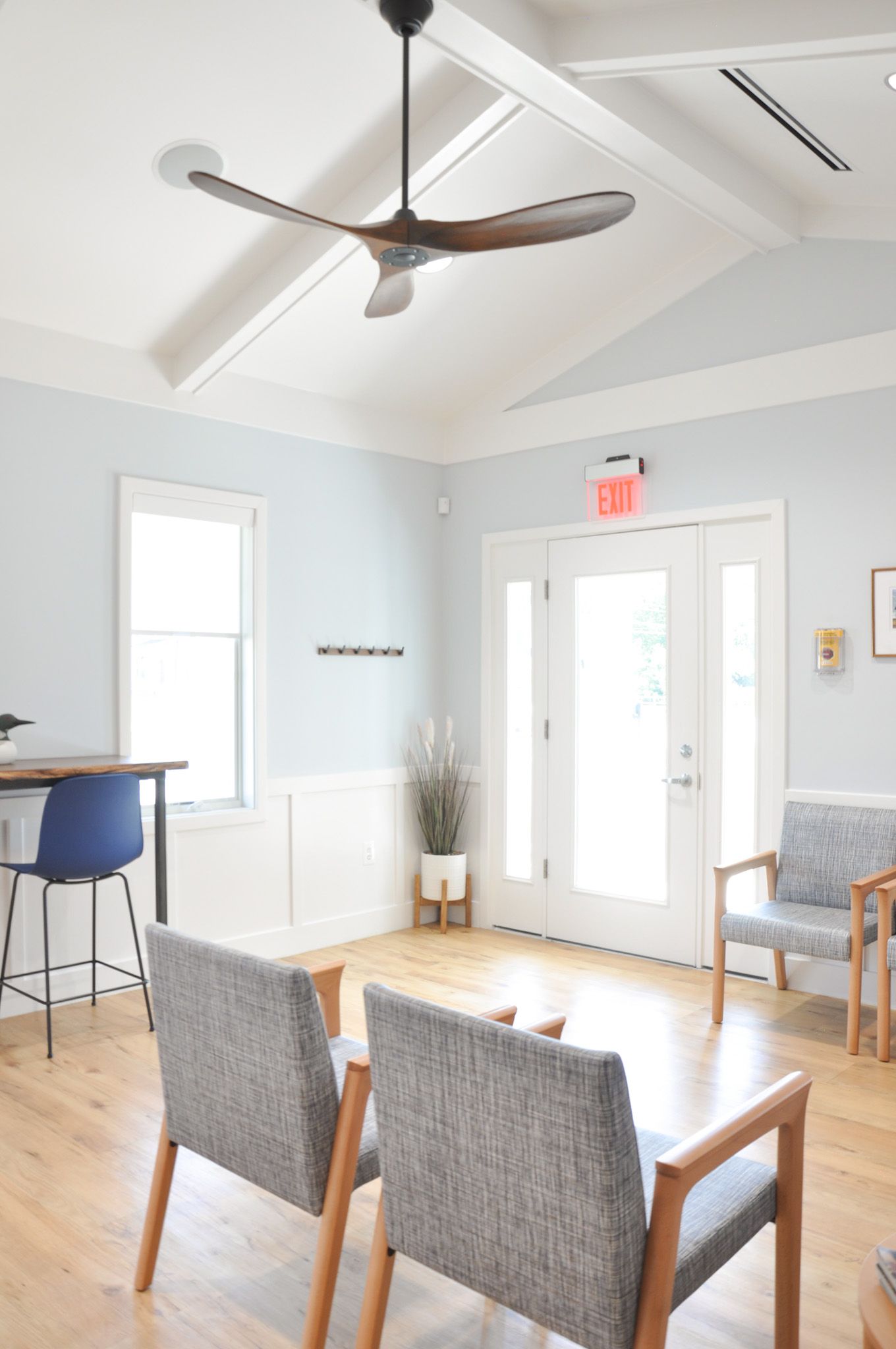 Waiting room with two gray chairs, a blue bar stool, and a ceiling fan. Light blue walls, white trim, and a wood floor.