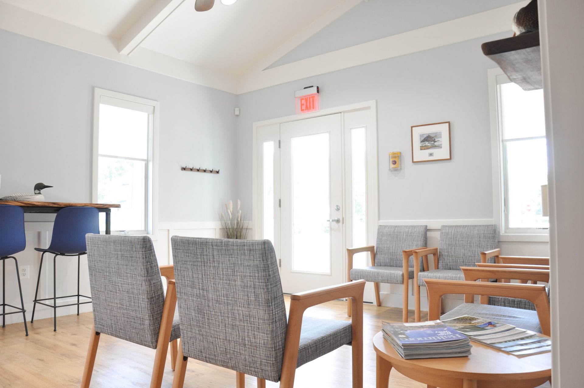 Waiting room with gray chairs, wooden tables, and large windows. Light blue walls and ceiling.