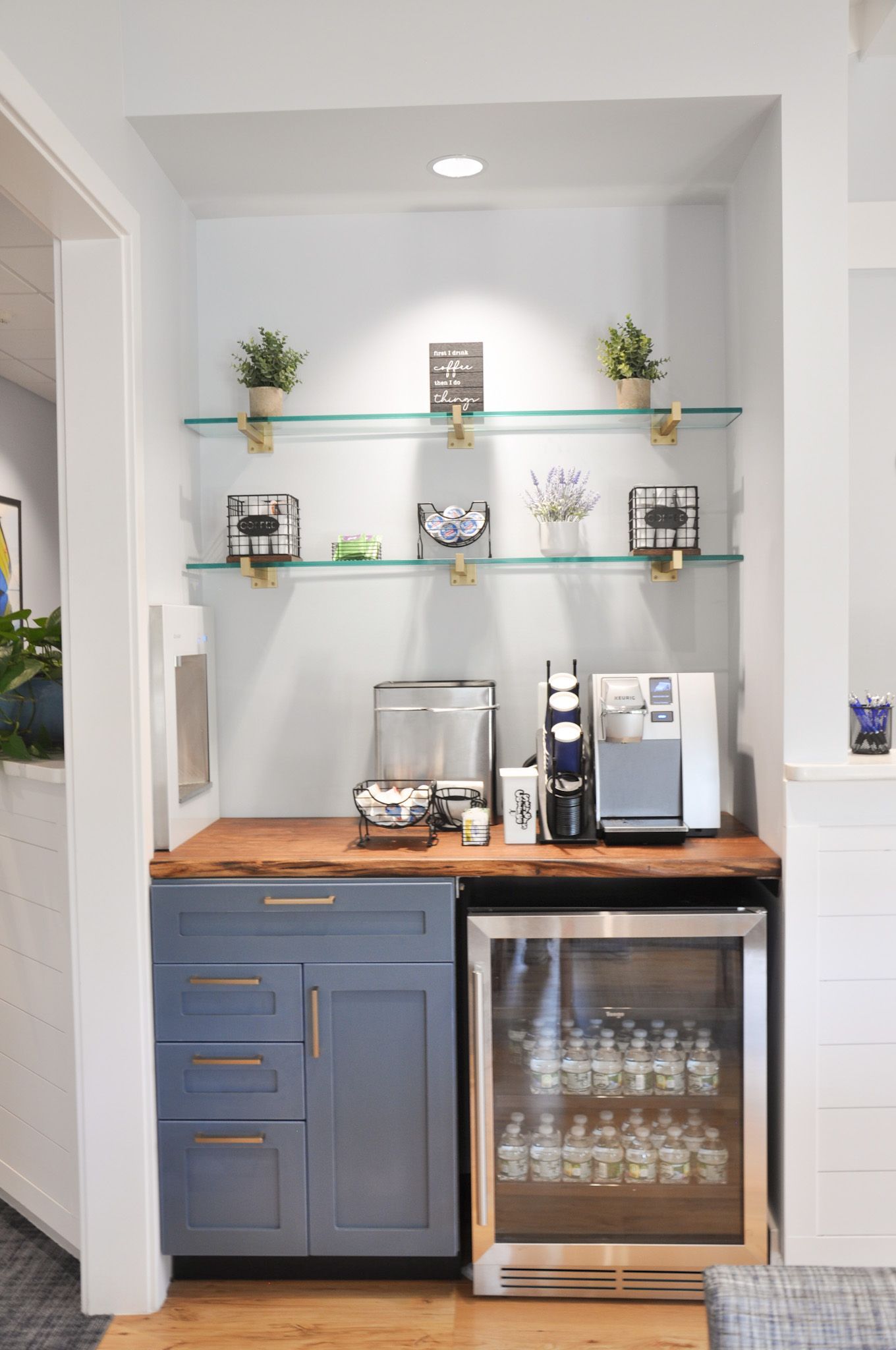 Built-in coffee station with cabinets, mini-fridge, and glass shelves displaying plants and decor.