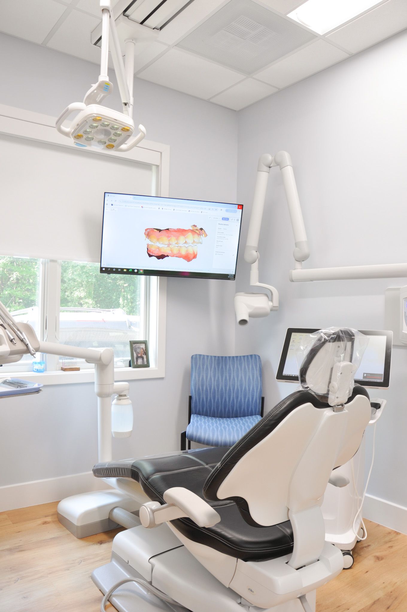 Dental examination room with chair, monitor showing teeth, equipment, and window.