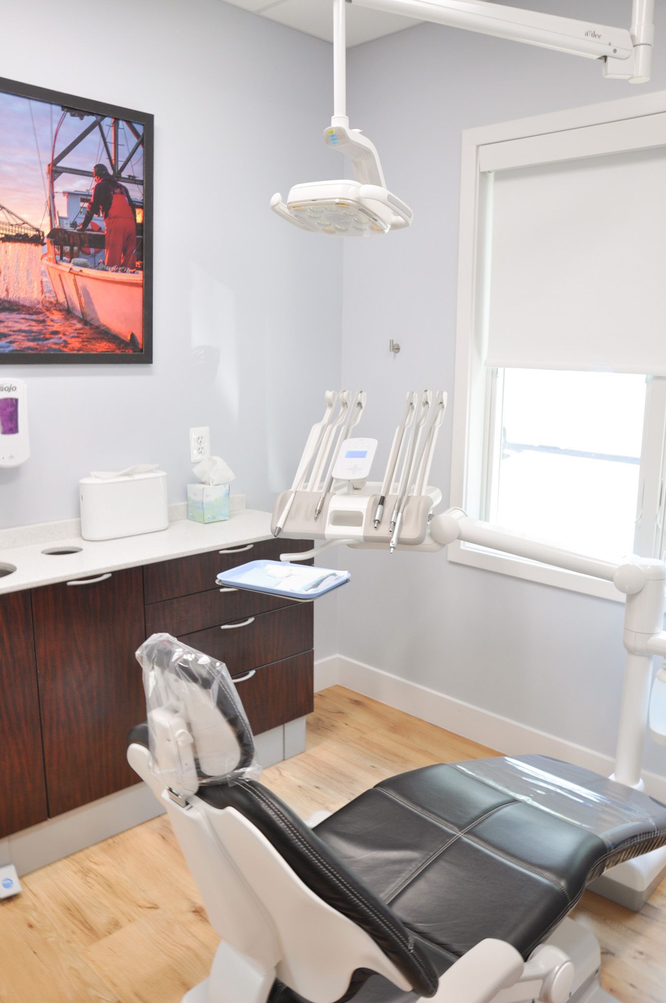 Dental exam room with chair, equipment, cabinets, window, and artwork.