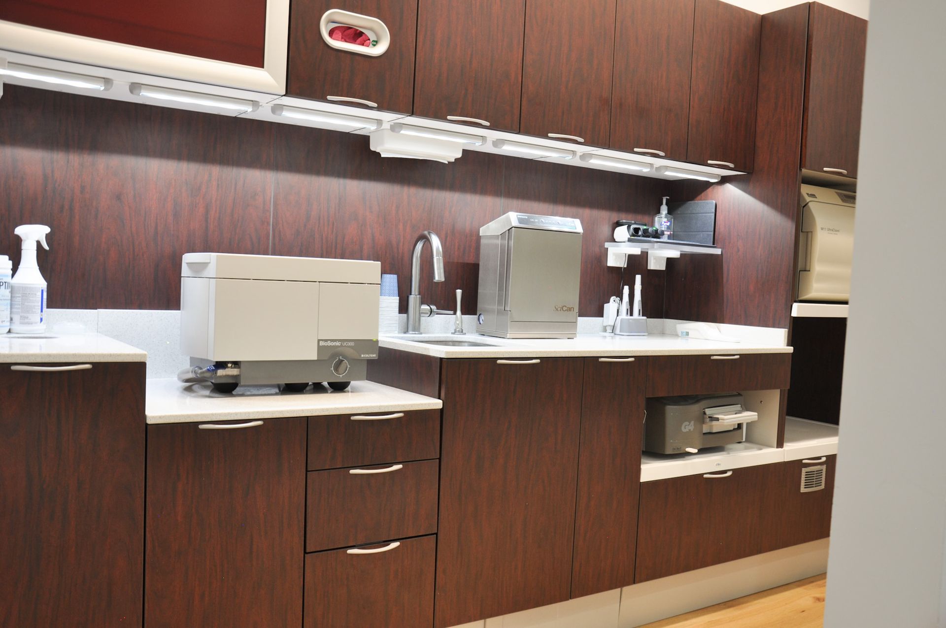 Dark wood cabinetry in a dental office. White countertop with sink, sterilizers, and storage, bright overhead lighting.