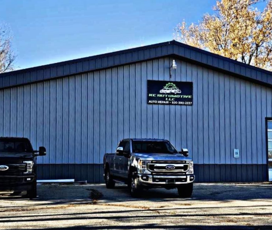 Gray building with a business sign, two pickup trucks parked in front. Autumn trees in the background.