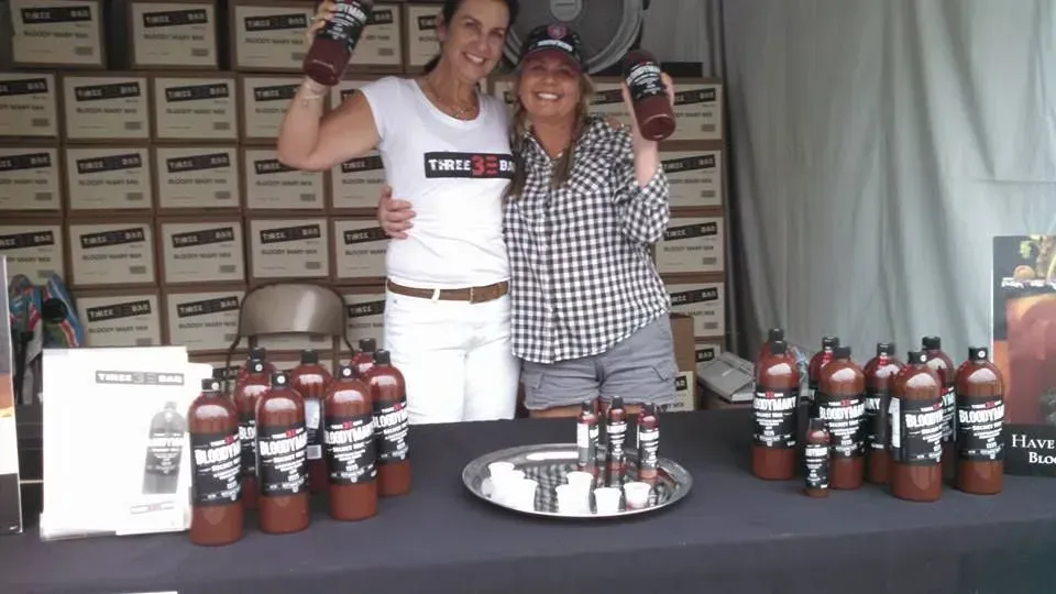 Two Women Standing in Front of a Table full Bloody Mary Mix Bottles