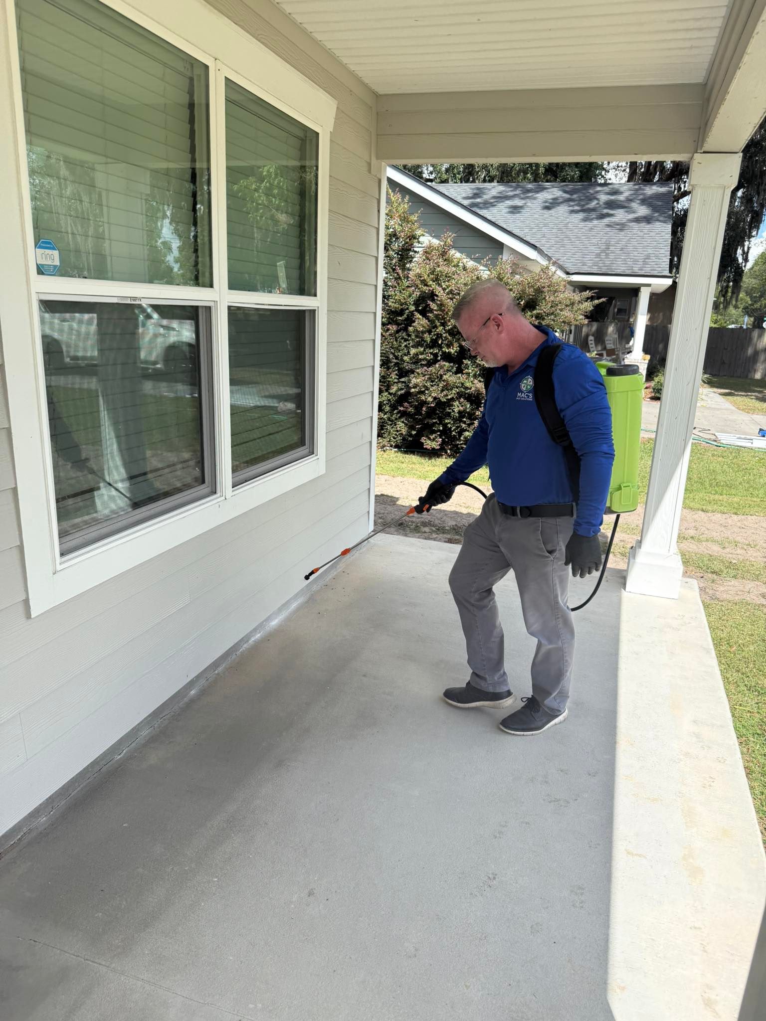Man sprays pesticide on a porch, wearing blue shirt, gray pants, with a green sprayer tank.