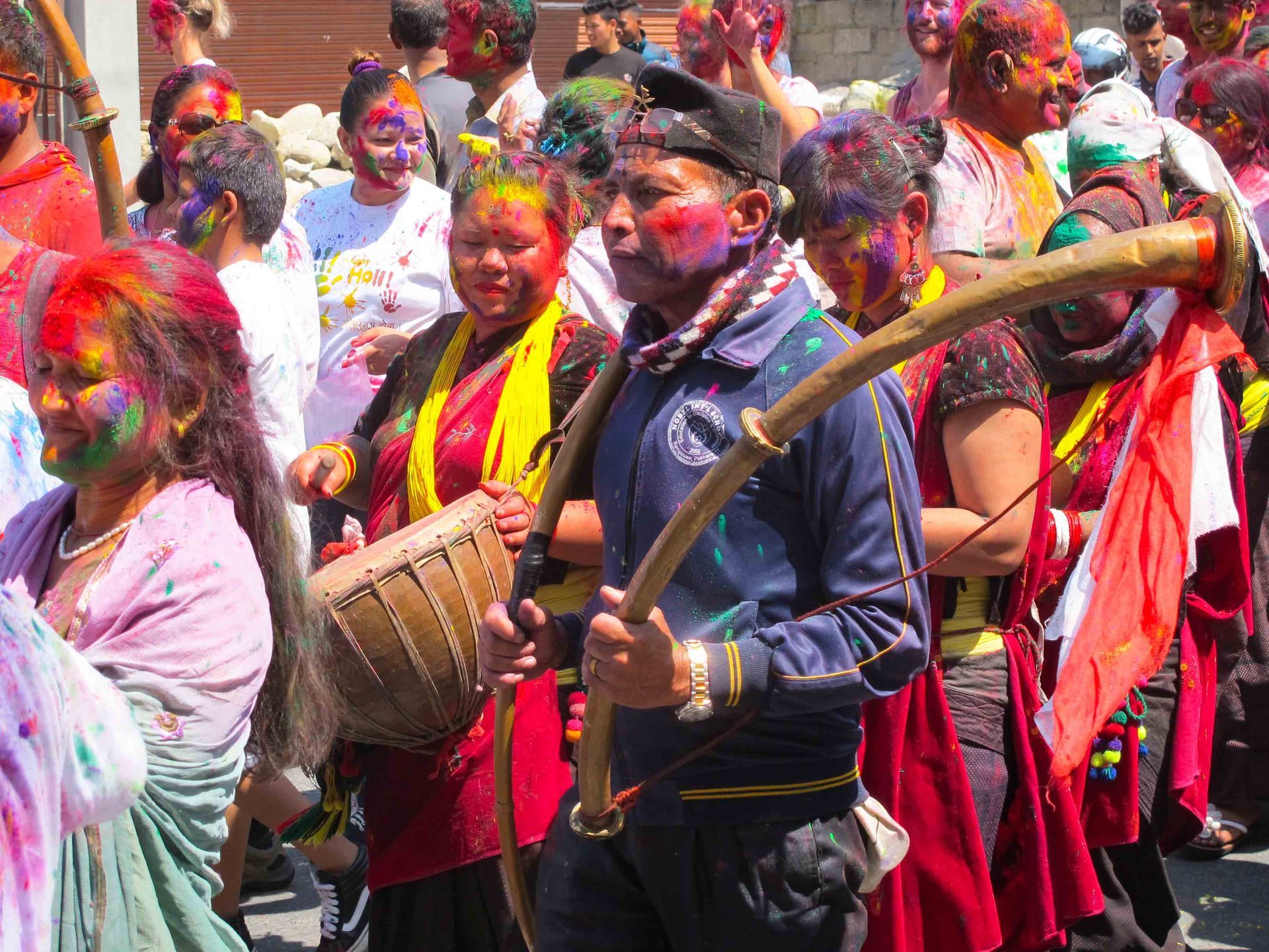 A lively crowd participates in the Holi festival in Pokhara, playing musical instruments amidst the colorful chaos.