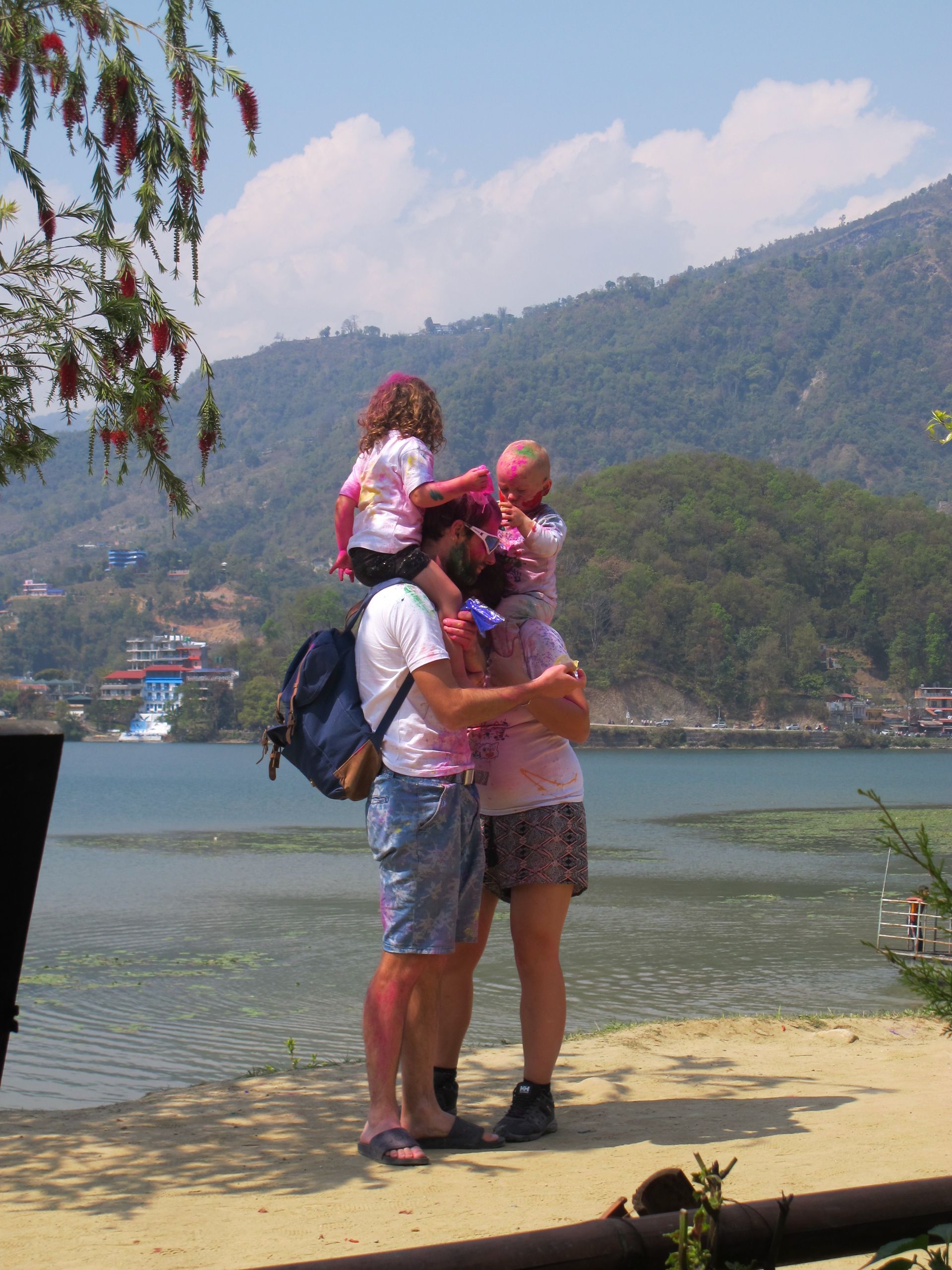 Family celebrating Holi in Nepal, covered in vibrant colors by a lakeside, enjoying the festive atmosphere.