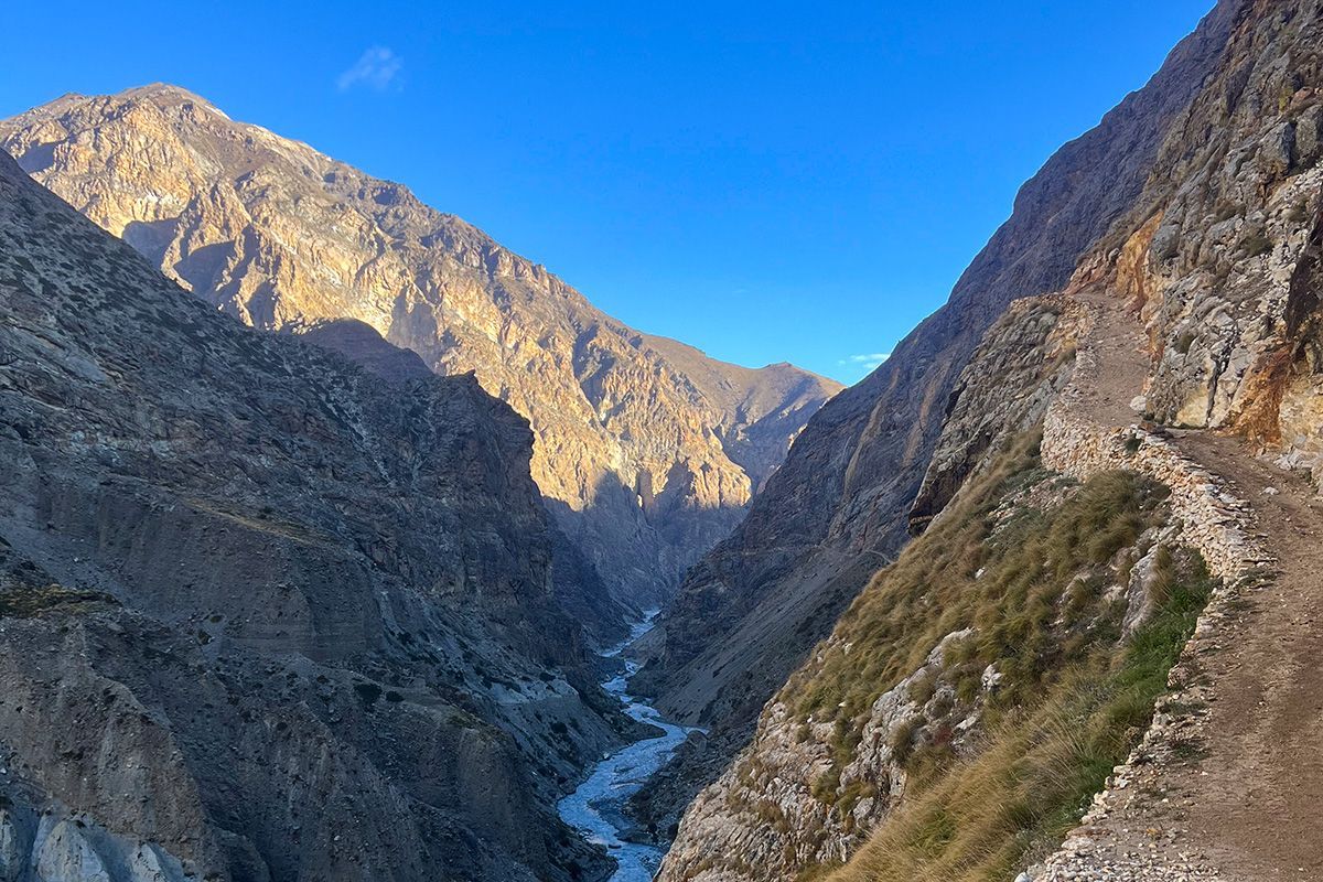 A rugged trail winding through the deep, dramatic Nar Phu Valley, flanked by towering rock faces and a river below.