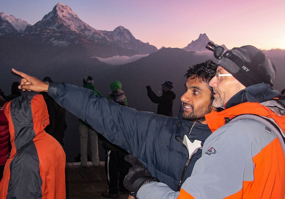 A guide and trekker in warm jackets admire the sunrise over the Himalayas, pointing towards the snow-capped peaks.